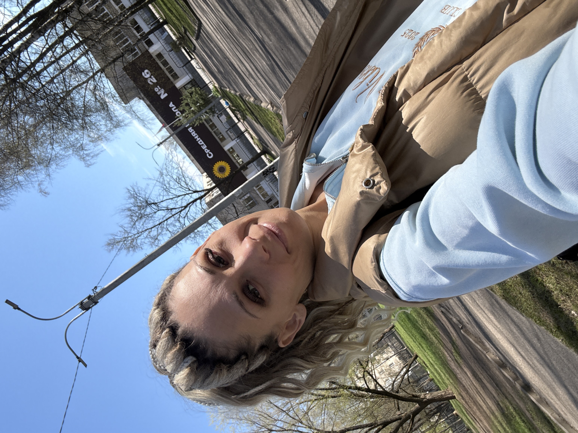 A woman stands outside on a sunny day with a clear blue sky. She is wearing a light blue sweatshirt and a brown jacket. Part of a building and a street can be seen in the background. The building has a sign that reads "СРЕДНЯЯ ШКОЛА № 26" with a sunflower underneath. This likely indicates that the image was taken in the city of Rusanovo, Russia where school number 26 is located.