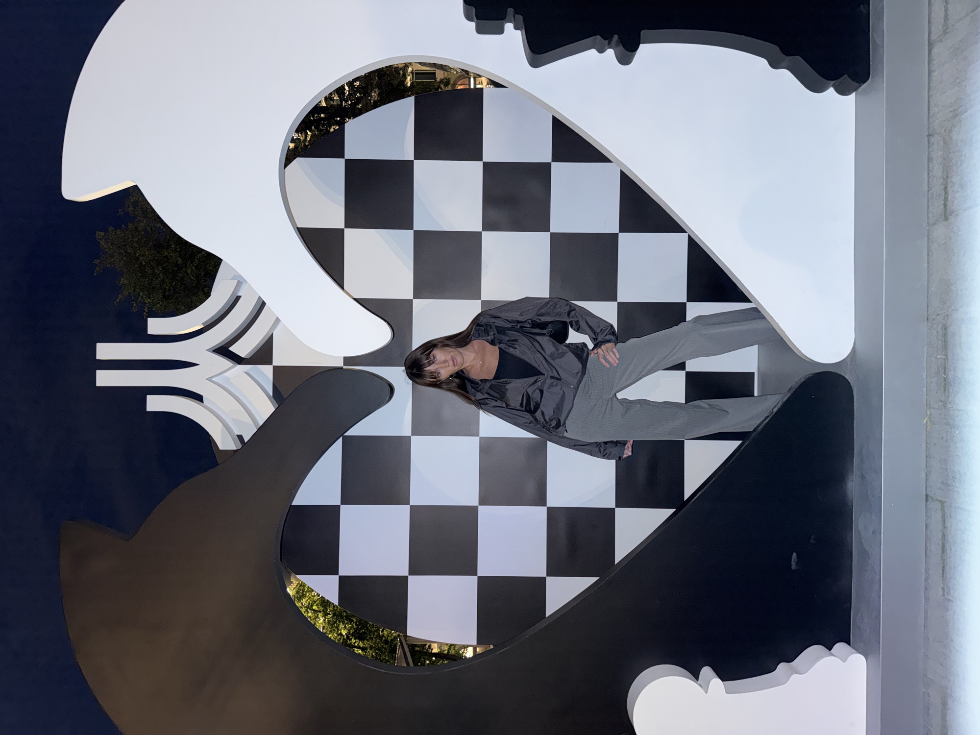 The image depicts a woman standing in front of a large, heart-shaped sculpture with a chessboard pattern inside. The sculpture is composed of two large chess pieces, one black and one white, forming the sides of the heart. The woman is wearing a dark-colored jacket, a black top, and grey striped pants. She is posing with one hand on her hip. The background appears to be a dark sky, with trees visible through openings in the sculpture. The sculpture is illuminated, and the scene suggests it is nighttime. This sculpture is located at the VDNKh (Exhibition of Achievements of National Economy) park in Moscow, Russia, specifically near the "Cosmonauts Alley" area. The sculpture is a temporary installation, often part of seasonal events or exhibitions.