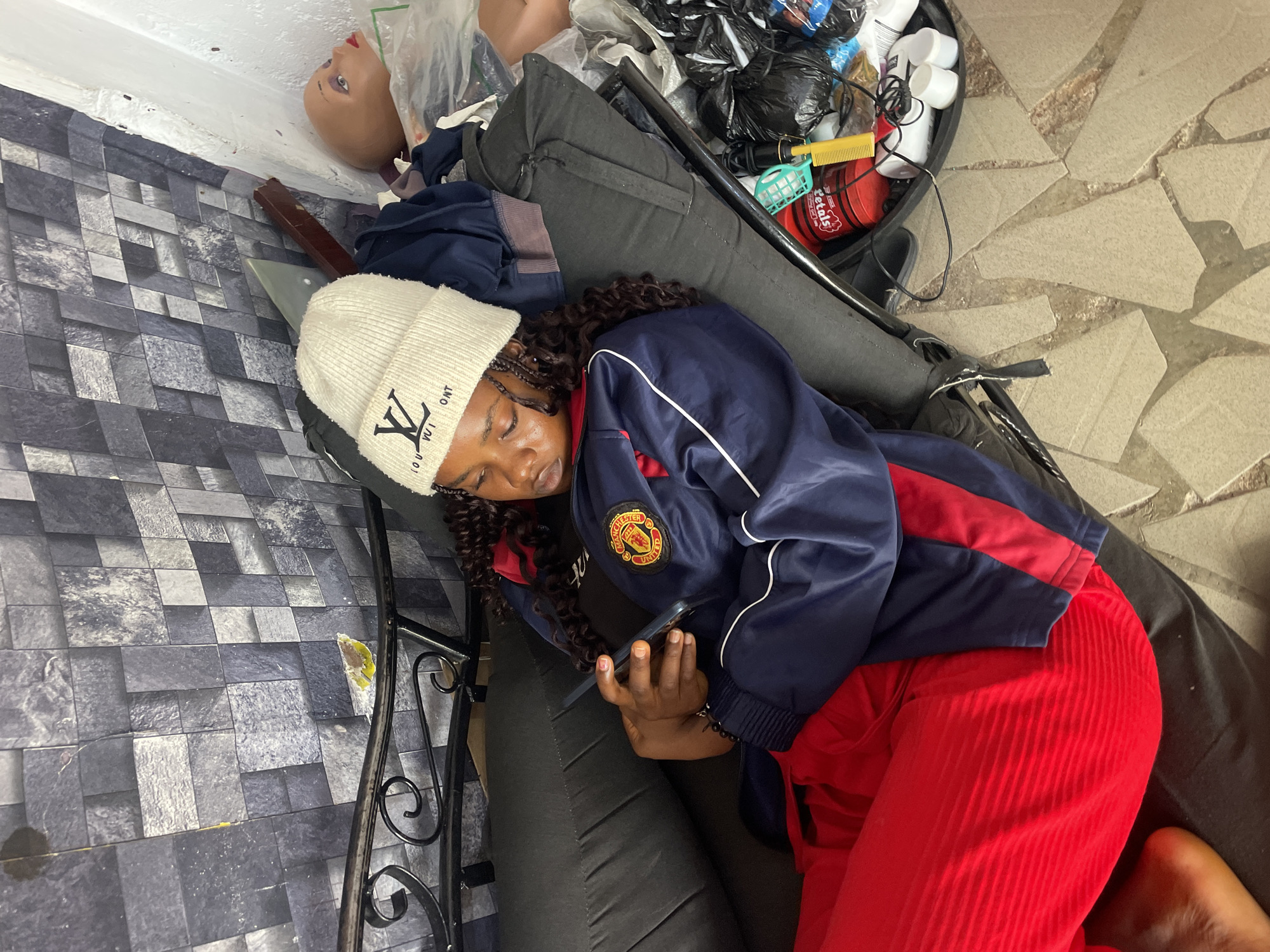 A person wearing a white beanie with a "LV" logo, a blue and red tracksuit jacket with a "Manchester United" logo on the left breast, and red pants is lying on a black padded cushion. The person is holding a phone in their hands. Next to the person, there is a bin filled with various items. To the left of the bin, there is a mannequin head. The environment includes a gray tiled wall and a light brown tiled floor. The location is likely indoors, based on the artificial lighting and the presence of domestic objects. The city is Jos, Nigeria.