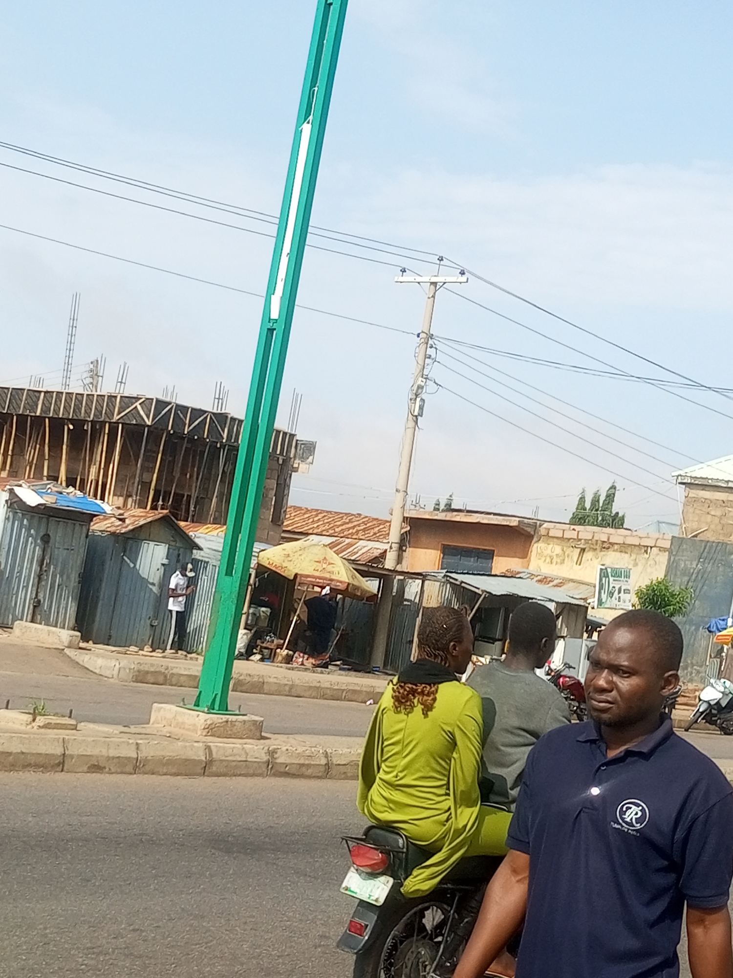 In Tempure, Nigeria, a man in a navy blue polo shirt with a white emblem stands in the foreground, facing slightly to the right, with a stern expression. Behind him, a woman wearing a bright green tunic is seated on a motorcycle, facing away from the camera. Another person, seen from the back and wearing a grey shirt, is also on the motorcycle. The motorcycle is parked on a paved road with a concrete curb. In the background, there are various structures, including buildings under construction with scaffolding, small metal shacks, and market stalls with umbrellas. Power lines and a tall, green pole are visible against a bright sky with some clouds. The scene appears to be a busy urban street.
