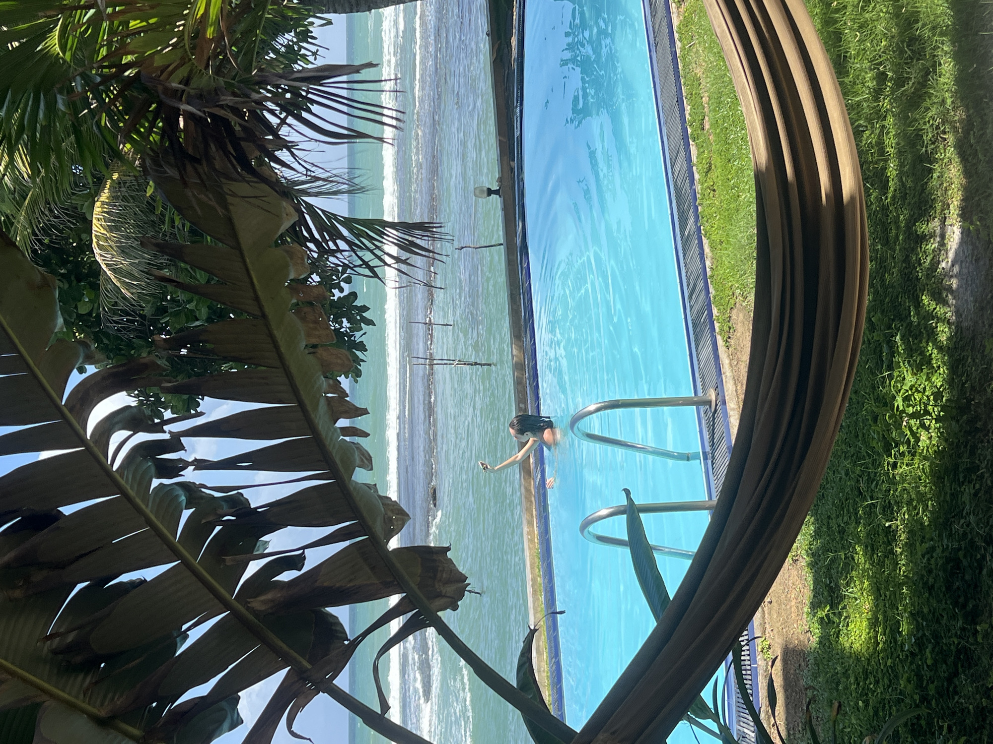 In Goviyapana, Sri Lanka, a woman is photographed in an outdoor swimming pool, holding a smartphone and appearing to take a selfie. The pool is filled with bright blue water and has metal ladder rungs visible. The pool area is surrounded by lush green grass. In the foreground, a hammock is stretched across the frame, obscuring some of the view. Beyond the pool, the ocean stretches to the horizon, with waves breaking and a clear blue sky overhead. Palm fronds frame the top of the image, adding to the tropical setting.
