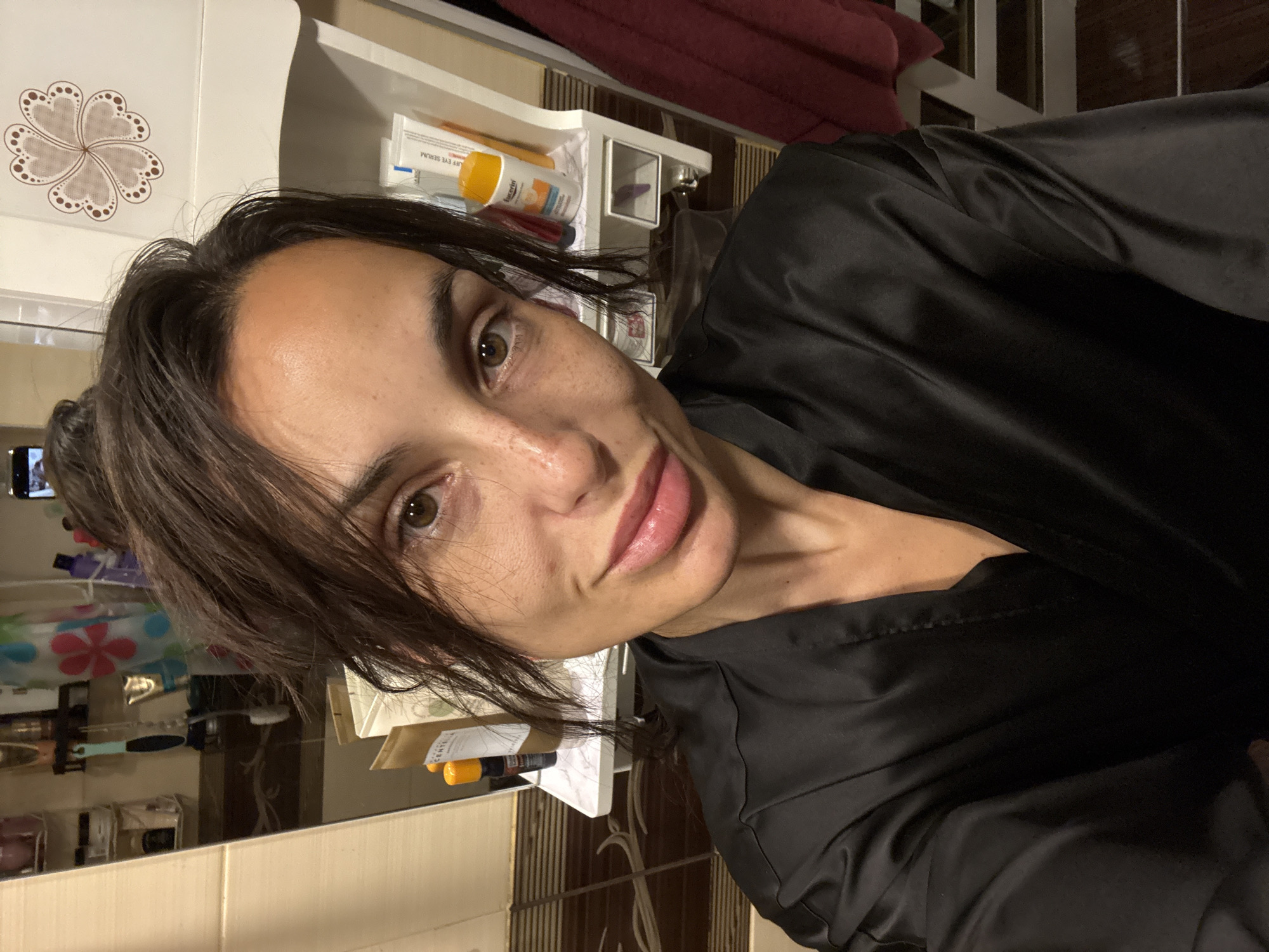 A woman is taking a selfie in a bathroom. She has dark brown hair pulled back in a bun and is wearing a black satin robe. She is looking at the camera with a slight smile. In the background, there are shelves with various toiletries and a mirror. The floor is tiled and there is a towel hanging on a rack. The overall impression is a casual, personal moment captured in a domestic setting. The location is not specified.