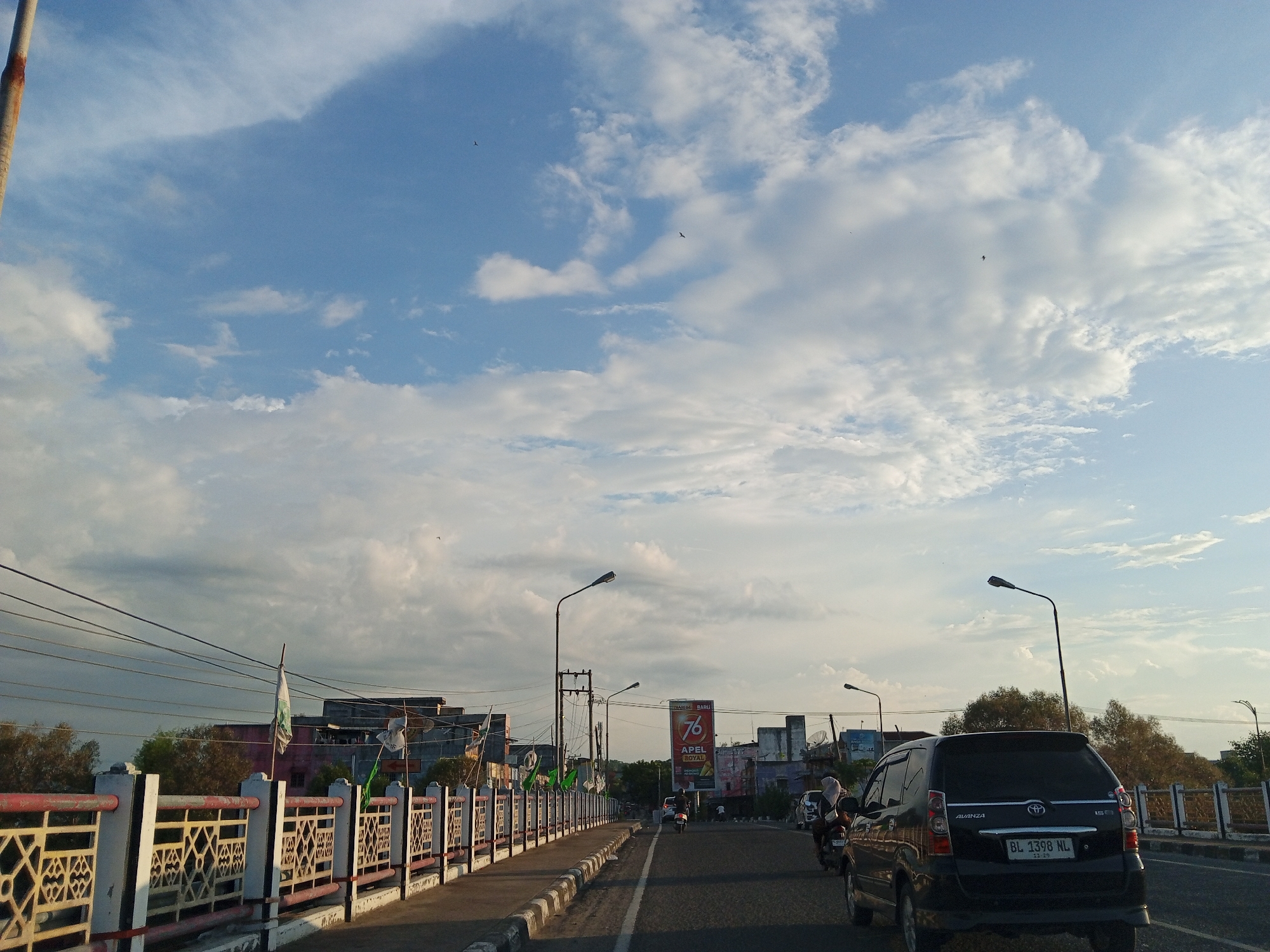 The image shows a road in Lhokseumawe, Indonesia, on a partly cloudy day. A black car, with license plate BL 1398 NL, is on the right side of the road. To the left is a bridge with a decorative railing. On the bridge are light poles and power lines. Across the bridge are buildings and a large sign reading "76 APEL ROYAL." Two birds are visible in the sky. A motorcycle is behind the car and moving in the same direction.