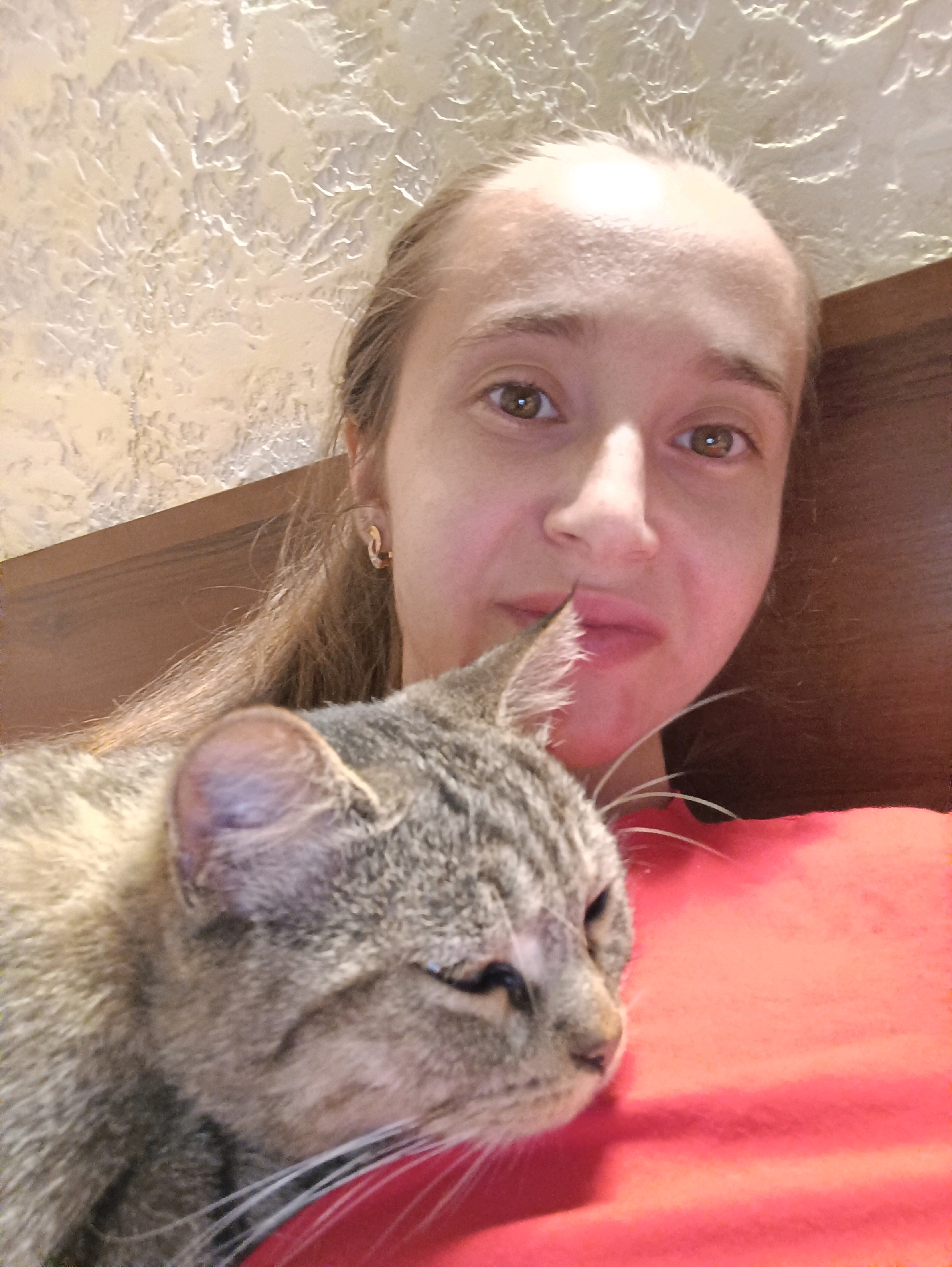 A medium shot captures a young woman with light brown hair pulled back and a tabby cat nestled against her. The woman is looking directly at the camera with a neutral expression, her eyes a warm brown. She is wearing a simple gold hoop earring in her visible ear. The cat, with its eyes partially closed, is resting on a vibrant red surface, likely a blanket or pillow. The background consists of a textured, light-colored wall and a wooden headboard. The scene suggests a moment of quiet companionship, possibly in a bedroom. The image does not provide any visual clues to indicate a specific location in Vypasne, Ukraine.