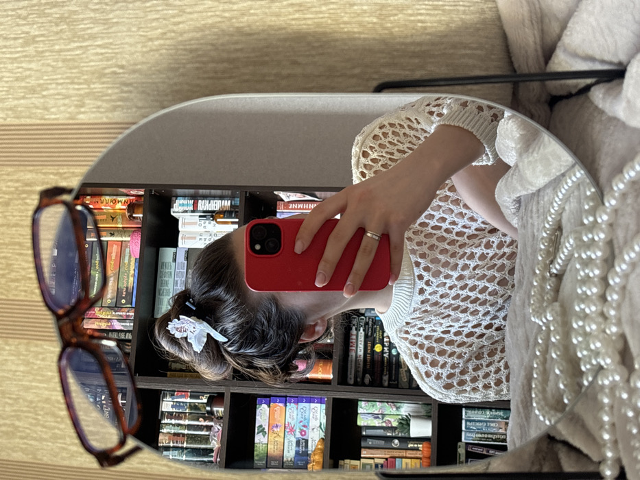The image depicts a mirror selfie taken indoors in Krasnodar, Russia. The central subject is an individual, presumably female, with dark brown hair styled in an updo secured with a white, possibly floral, hair clip. This individual is holding a red smartphone, likely an iPhone due to the visible camera configuration, which obscures their face. A two-tone metallic ring is worn on the left ring finger, and light pink nail polish is visible. The individual is wearing a light-colored, open-knit or crocheted long-sleeved top with a ribbed collar.
Reflected in the mirror behind the individual is a dark wooden bookshelf filled with numerous books, some with colorful spines, and various small objects including a miniature plant in a white pot, an orange decorative pumpkin, and several small bottles. The mirror itself is framed in black and appears to be placed on a soft, light-colored surface, possibly a blanket or rug, on which a strand of white pearls is laid out in the foreground. Resting on the upper edge of the mirror's frame are a pair of brown-framed eyeglasses. The wall visible outside the mirror's reflection features light brown, vertically striped wallpaper.