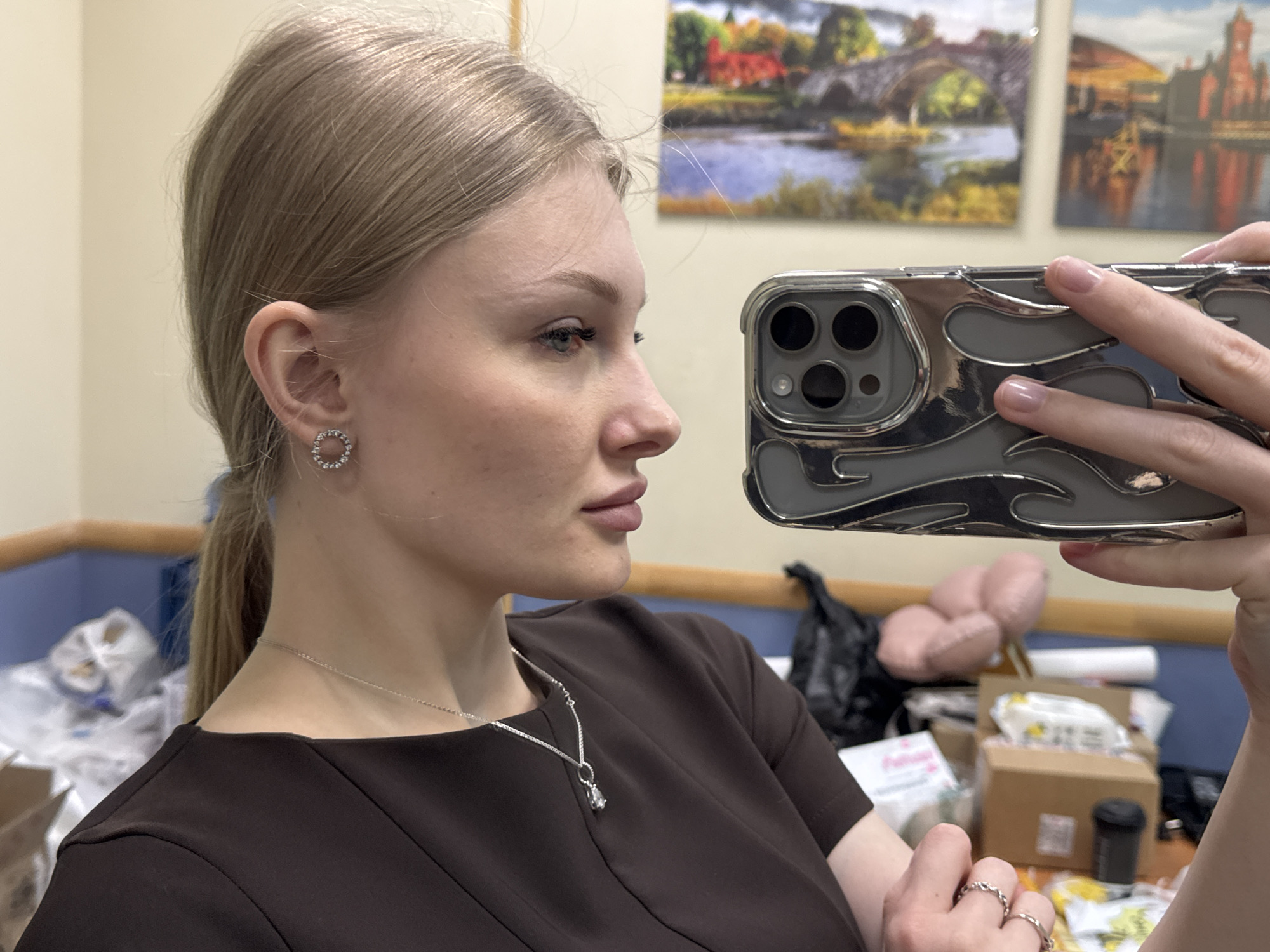 A woman takes a selfie. She is fair-skinned with blonde hair pulled back in a ponytail. She is wearing round stud earrings, a silver necklace with a teardrop pendant, a dark brown dress, and a ring. Her phone is silver with a decorative case. The background includes two framed images on a wall depicting a landscape with a river and a bridge. The scene is a cluttered office with boxes, a black garbage bag, and other objects. There is no indication of Perm, Russia.