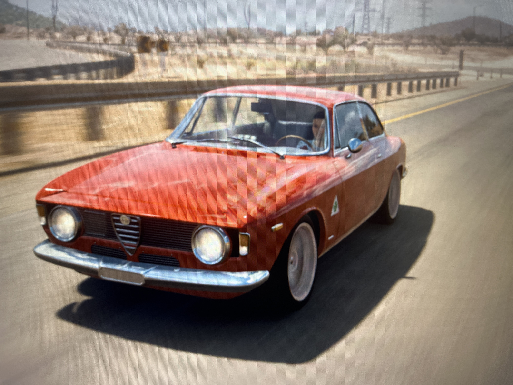 A red, classic Alfa Romeo coupe is driving on a paved road with a slight curve to the left. The car is the primary subject and is in motion, indicated by motion blur on the background. The driver, a person with light-colored hair, is visible through the windshield, with their right hand on the steering wheel. The environment appears to be arid with sparse vegetation and a guardrail lining the side of the road. In the distance, there are power lines and hills. The lighting suggests it is daytime. The scene does not contain any elements that would specifically place it in the city of Varna, Bulgaria.