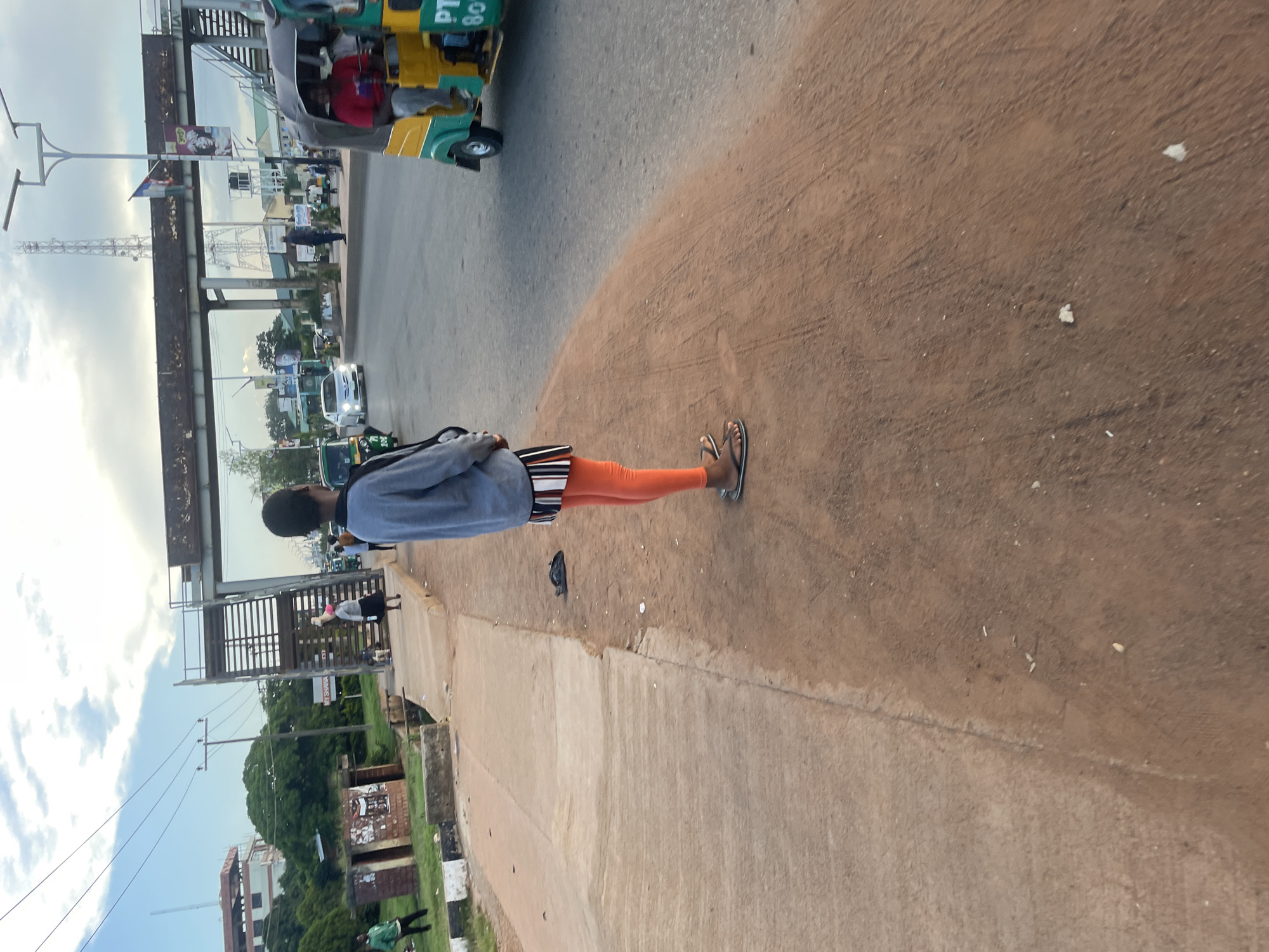 The image depicts a street scene in Jos, Nigeria, with a focus on a person standing on a dirt path next to a paved road. The person, viewed from the back, is wearing an oversized grey sweatshirt, a striped skirt with black, white, and orange bands, and orange leggings, along with black flip-flops. In the background, traffic is visible, including a yellow and green auto-rickshaw with the lettering "PTY" and the number "80" on its side, and a white car. A pedestrian bridge with metal stairs spans the road. On the left side of the image, a low-lying building with brick and concrete elements and a damaged roof is visible, along with some greenery and a smaller structure. The sky above is partly cloudy with a mix of blue and white. The overall scene suggests an urban or semi-urban environment with active transportation and pedestrian infrastructure.