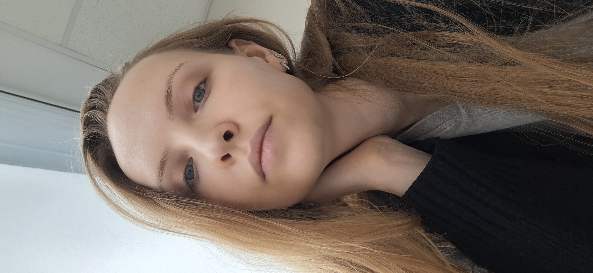 A young woman with light skin, blue eyes, and long light brown hair is lying on her side. Her head is resting on her hand. She is wearing a black knitted sweater over a grey top. The background shows a white wall or ceiling. It is not possible to identify the location as Chaikovsky, Russia from the image alone.