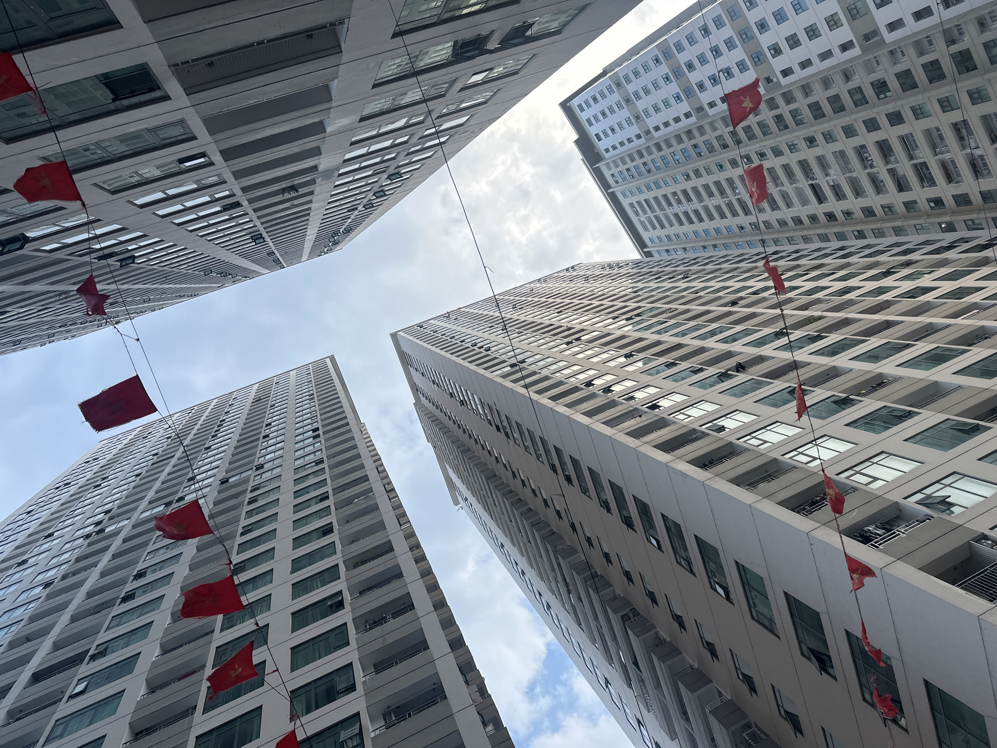 The photograph captures a low-angle view of four high-rise buildings in Bac Nha Trang Ward, Vietnam. The buildings, constructed with a modern architectural style, feature repeated patterns of windows and balconies. Strung between the buildings are rows of small red flags, presumably for a celebratory event. The sky is overcast with a mix of white and gray clouds peeking between the towers. There are no visible people in the image.