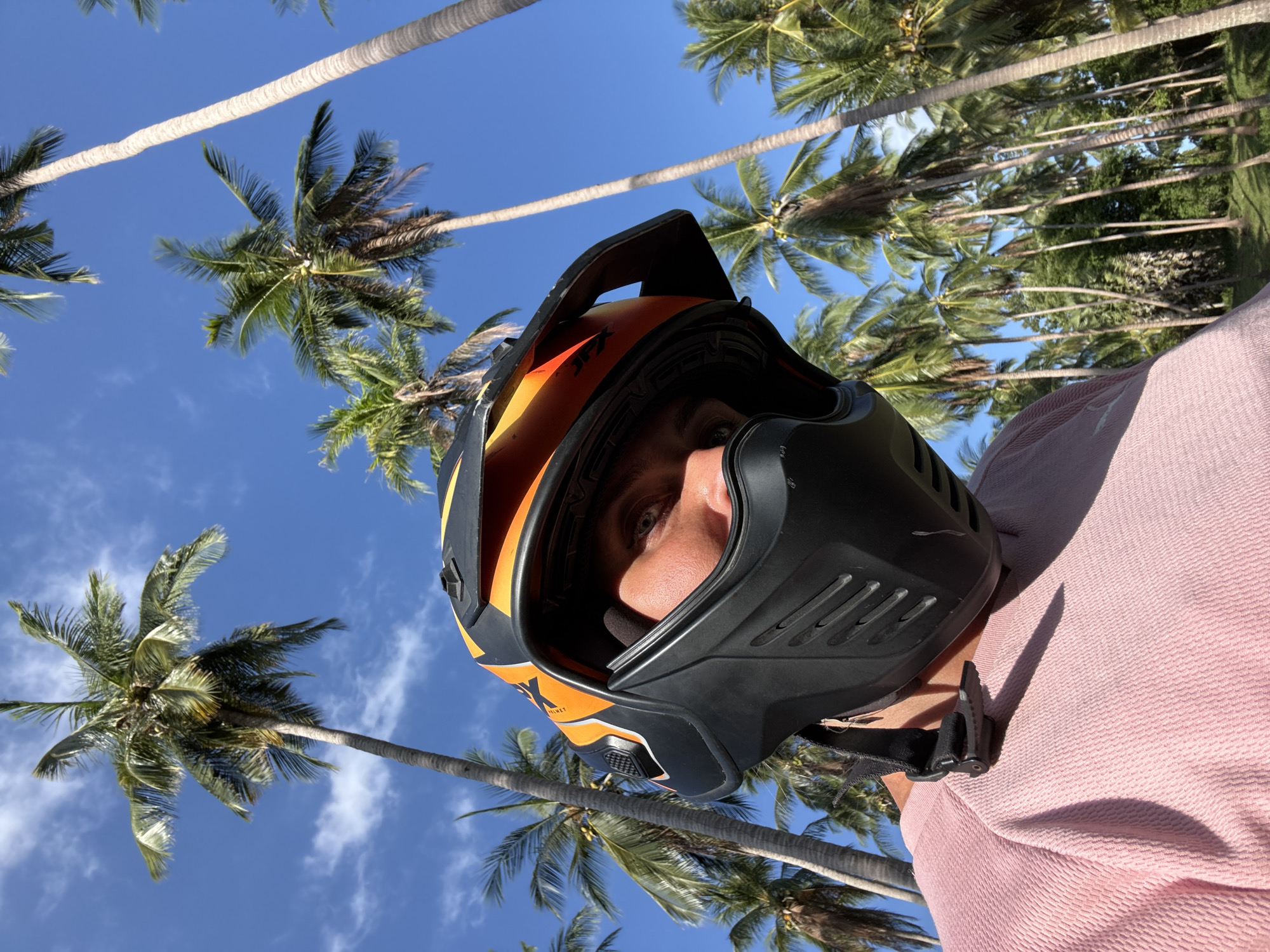 A low-angle, close-up, selfie-style shot shows a person wearing a black and orange motorcycle helmet, with a portion of their face visible. The person is wearing a pink long-sleeved shirt. The background is a vibrant blue sky with scattered white clouds, filled with numerous tall palm trees. The scene appears to be in a tropical location, consistent with Ko Samui District, Thailand. The lighting suggests it is daytime.