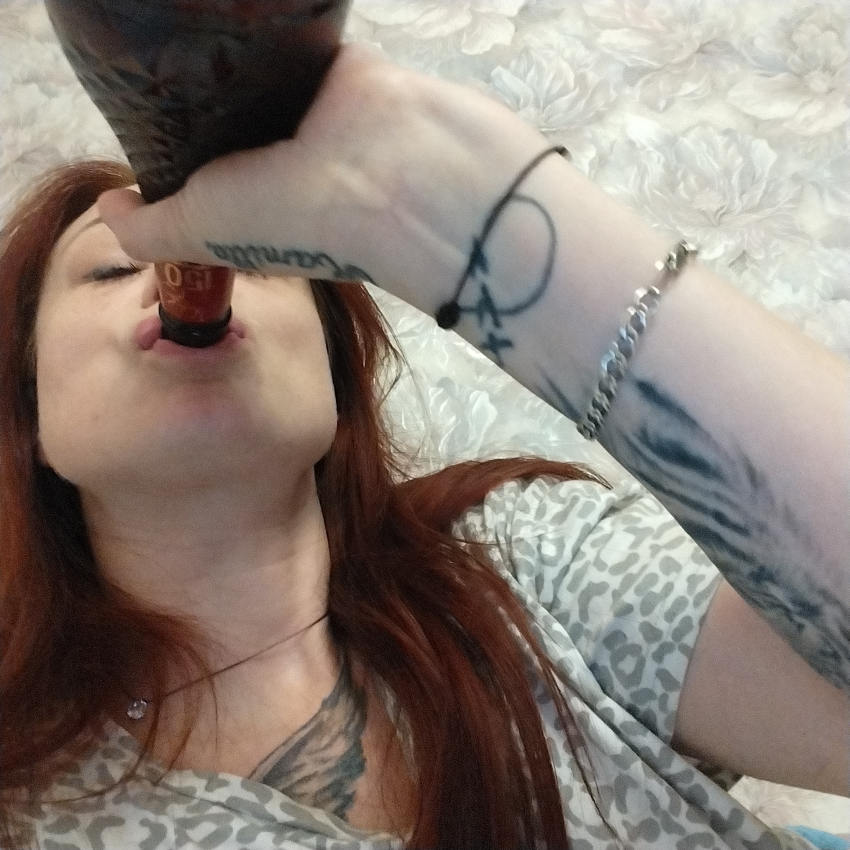 A close-up, indoor shot shows a woman with reddish-brown hair drinking from a dark bottle. She is lying down, and the camera is positioned above her, looking down. The woman's eyes are closed, and her lips are slightly parted as she drinks. She is wearing a light-colored, patterned shirt. The background is a light-colored, textured surface, possibly a blanket or bedspread. Her right arm is visible, with tattoos on her forearm and wrist, and she is wearing a silver bracelet. A thin necklace with a small, clear pendant is visible around her neck. The scene appears to be in a domestic setting, as indicated by the soft furnishings in the background.