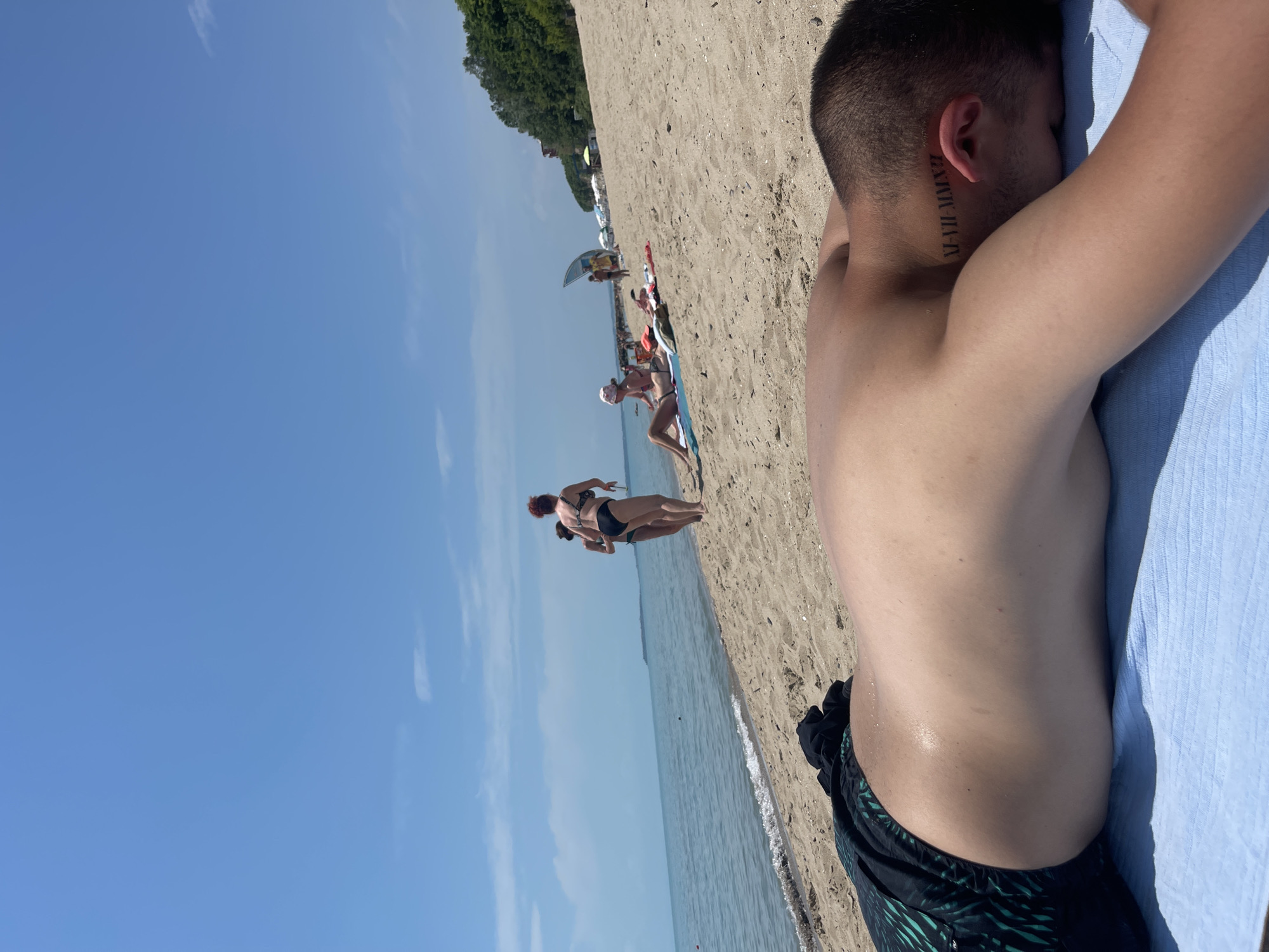 This image depicts a bright and sunny day at a beach, likely in Sveti Vlas, Bulgaria. The primary subject in the foreground, seen from a low angle on the right, is a person lying on their stomach on a light blue towel. The person's bare back, short dark hair, and dark swim shorts with a tropical pattern are visible. A significant detail is a Roman numeral tattoo "XI-VII-MMXII" on the back of their neck, near the left ear.
The scene extends along a sandy beach bordering calm, blue water under a clear, expansive blue sky with a few faint clouds. In the midground, several other beachgoers are visible. Two women stand near the water's edge, one wearing a dark bikini and facing away from the camera. Further down the shoreline, more indistinct figures can be seen relaxing on towels, some near the water, and beach amenities such as umbrellas and sunbeds are present. A distant treelined hill or coastline forms the horizon. The overall impression is one of a peaceful and leisurely day spent enjoying the sun and sea.
