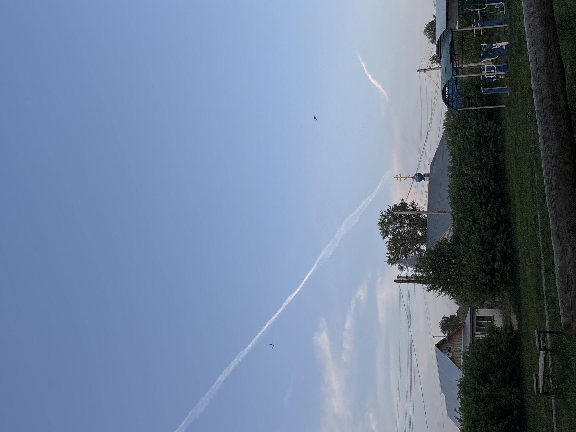 The image shows a sky with contrails and birds flying above a small village. The location is possibly in Фоки, Russia. The foreground contains trees and houses. A sports structure is visible. Utility poles and lines are also present.