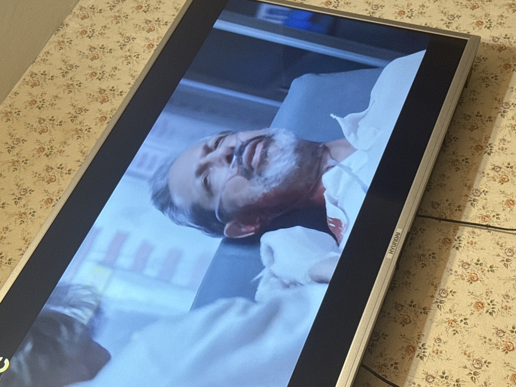 The image shows a Hyundai flat-screen television displaying a scene with a man lying in what appears to be a hospital bed. He has a white beard, an oxygen tube in his nostrils, and appears to be unconscious or ill. He is covered with a white sheet. The background shows medical equipment and a hospital-like setting. The television is situated in a room with a floral patterned wallpaper.
There is no information in the image that would allow identification of a specific location in Mytishchi, Russia.