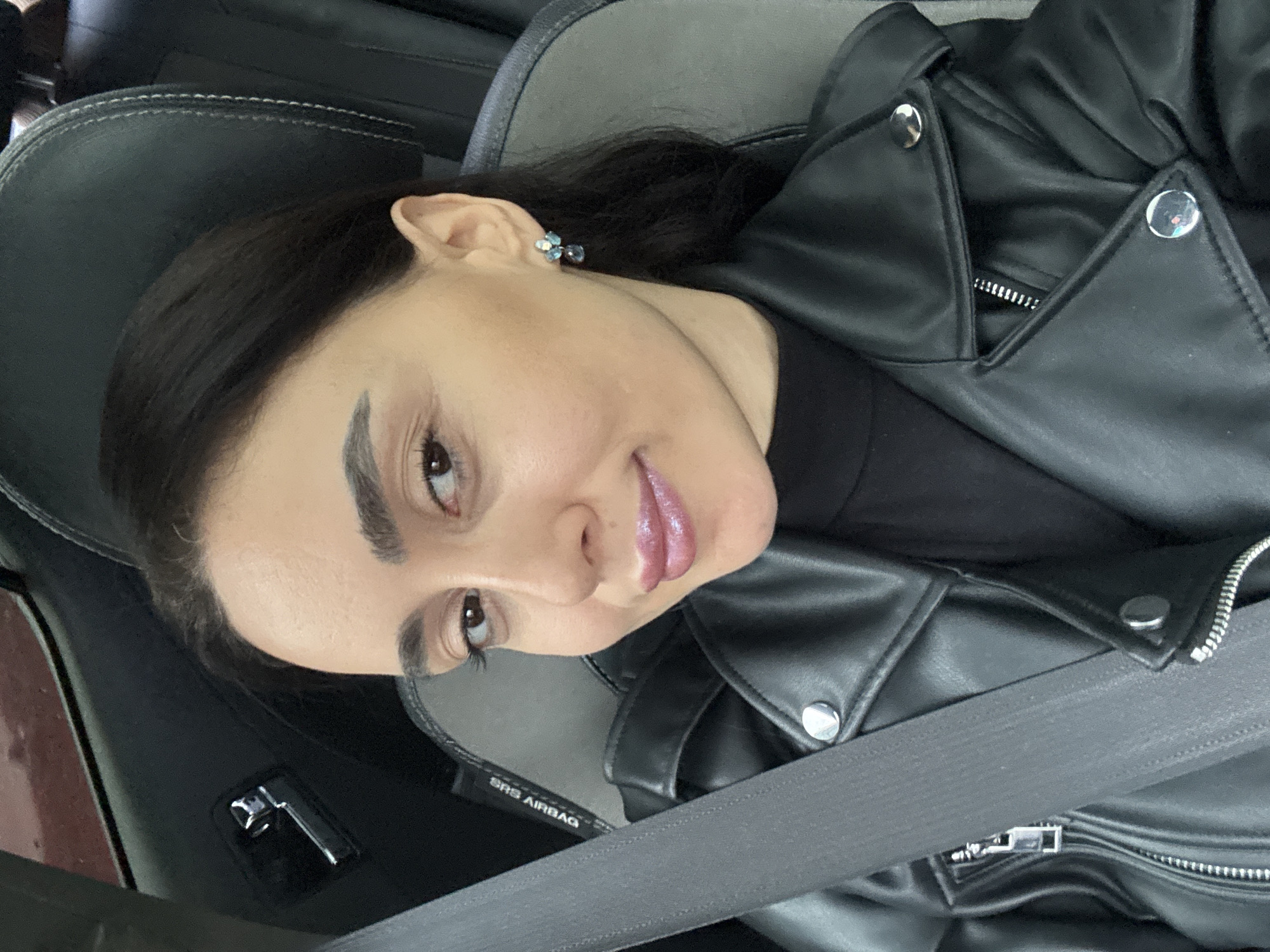 A female subject is observed in the front passenger seat of a vehicle in Tbilisi, Georgia. Her upper torso and head are visible, angled towards the camera. She is wearing a black leather jacket over a black garment, secured by a grey seatbelt across her left shoulder and chest. The subject has dark hair, brown eyes, and is wearing makeup including pink glossy lipstick. A multi-stone blue earring is visible on her right ear. The vehicle interior features dark grey upholstery with white stitching on the headrest, and a chrome door handle on the driver's side door panel. An "SRS AIRBAG" label is visible near the seatbelt mount.
