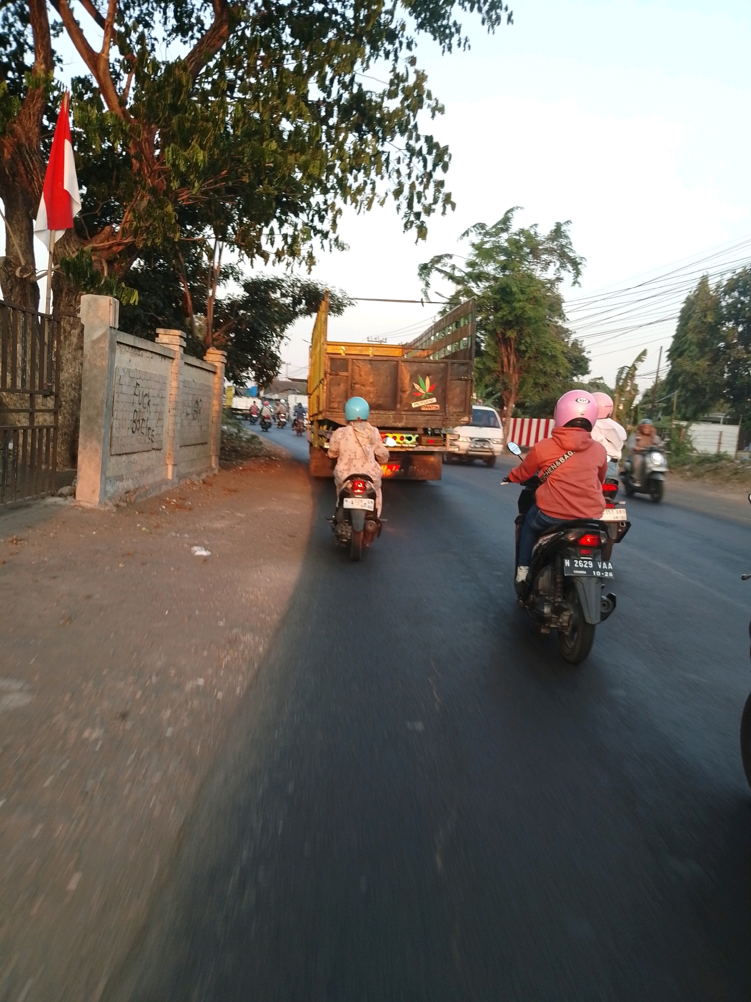 The image depicts a street scene in Pasuruan, Indonesia, with a focus on vehicles and their occupants. In the foreground, a person wearing a pink helmet and an orange hoodie rides a motorcycle. The motorcycle's license plate reads "N 2629 VAA 10-26". Another person on a motorcycle with a light blue helmet is visible further ahead, to the left. Behind them, a large yellow truck with "PUTRA SUMANTRI" written on its side is in motion. A white van is positioned behind the truck. The road is paved, with a dirt shoulder on the left side of the image. A concrete wall with graffiti, including the words "FUCK BOTICE", is visible along the shoulder. Trees line the side of the road, and a red and white Indonesian flag is partially visible hanging from a tree. The sky is clear and bright, suggesting it is daytime. The image appears to be captured from a moving vehicle, as indicated by the motion blur.