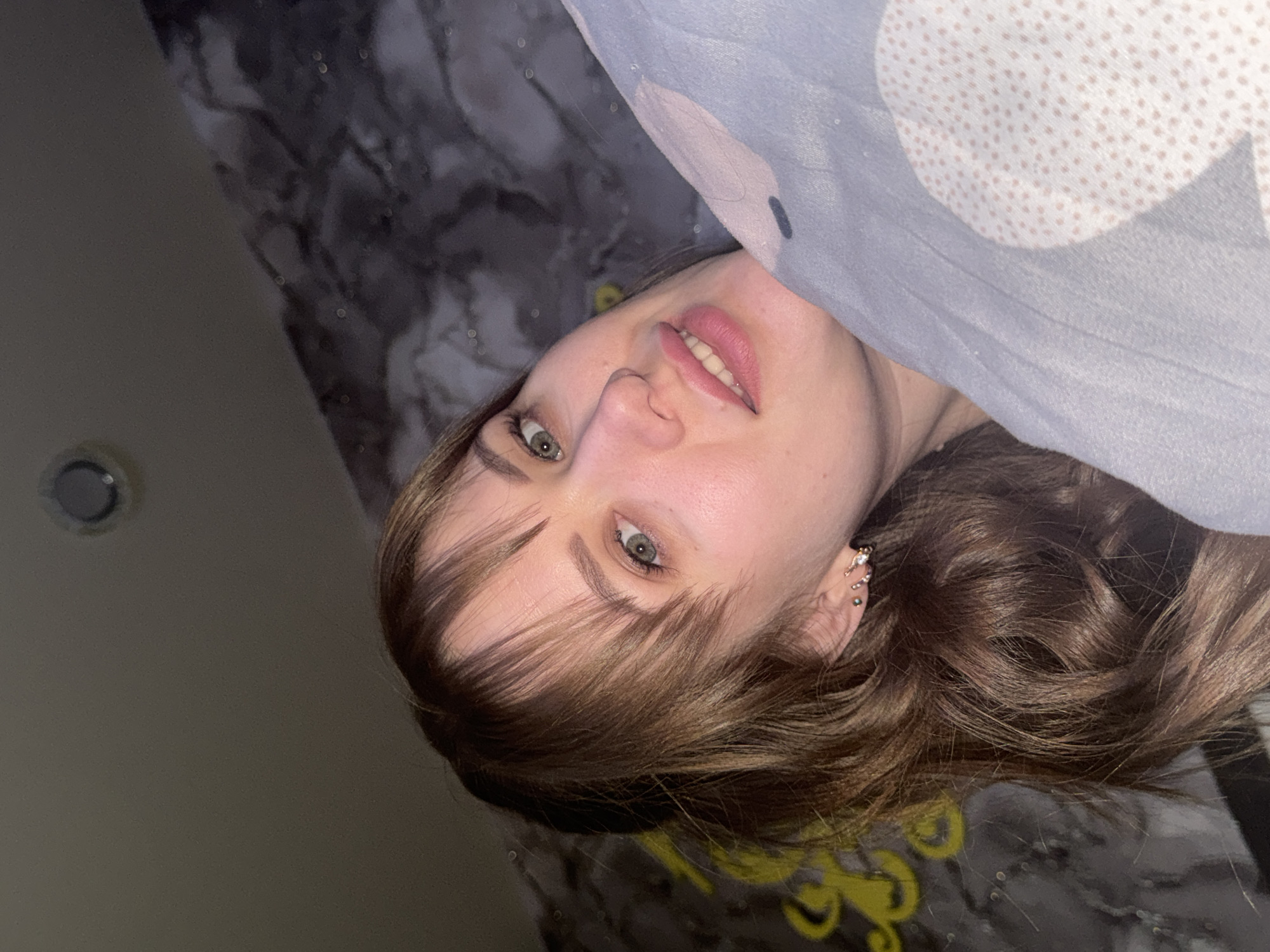 A person is lying down, partially covered by a grey and white polka-dotted fabric, possibly a blanket. They have fair skin, light brown hair with bangs, and green eyes. They are wearing earrings. The background consists of a grey patterned surface, likely a bed or bedding, and a dark grey wall with a visible light fixture. There is no indication of Chaikovsky, Russia.