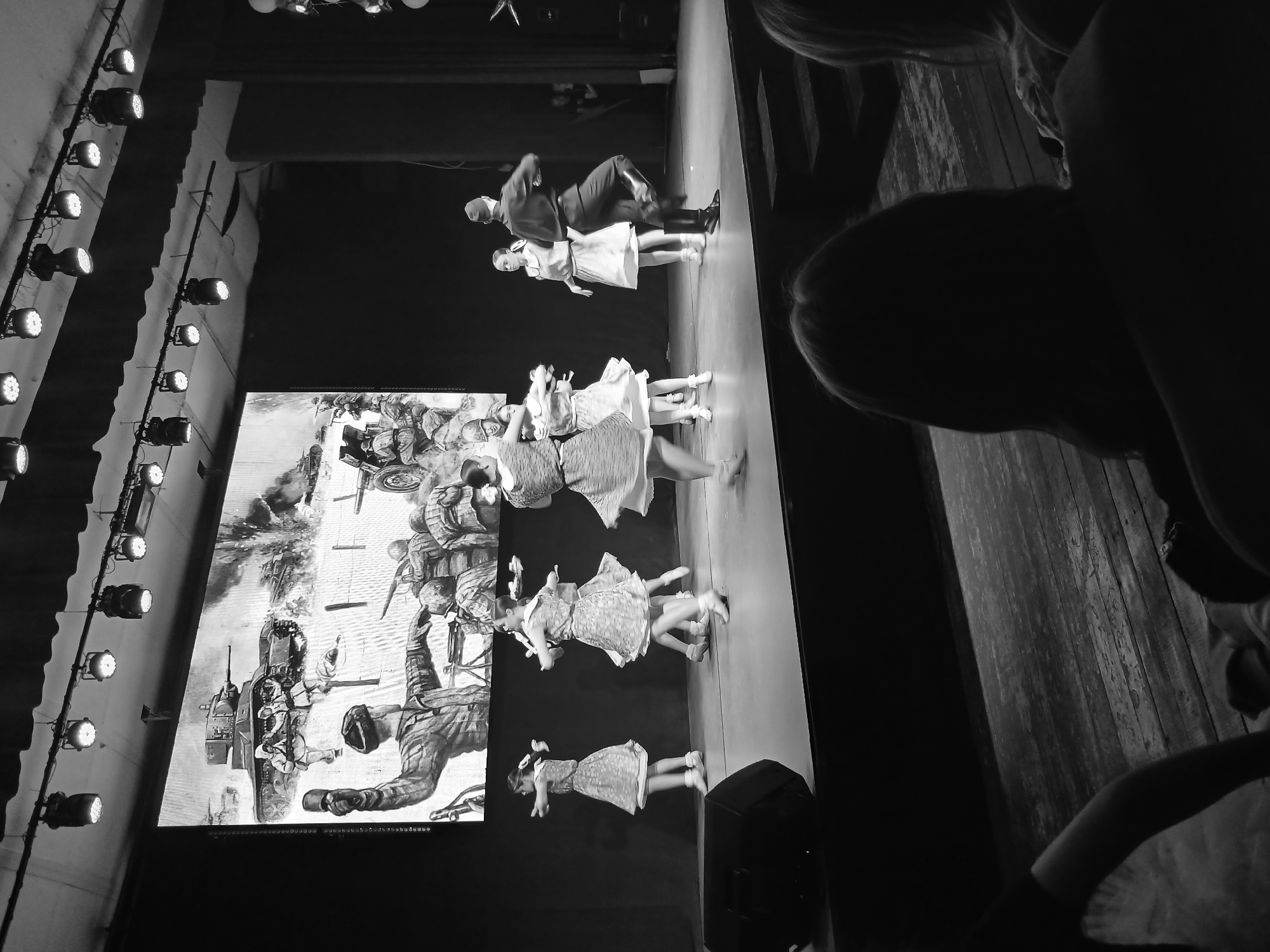 The image shows a performance taking place on a stage, likely in Votkinsk, Russia, judging by the context. On the left, a large screen displays a monochrome image of a war scene, possibly depicting World War II, complete with tanks and soldiers.
Several performers are on stage. There are multiple young girls in dresses, and one man and woman are in more formal attire. The man is mid-leap, suggesting a dance or theatrical performance.
The environment consists of stage lighting, a dark backdrop, and the edge of the audience's view in the foreground. The image is in black and white.