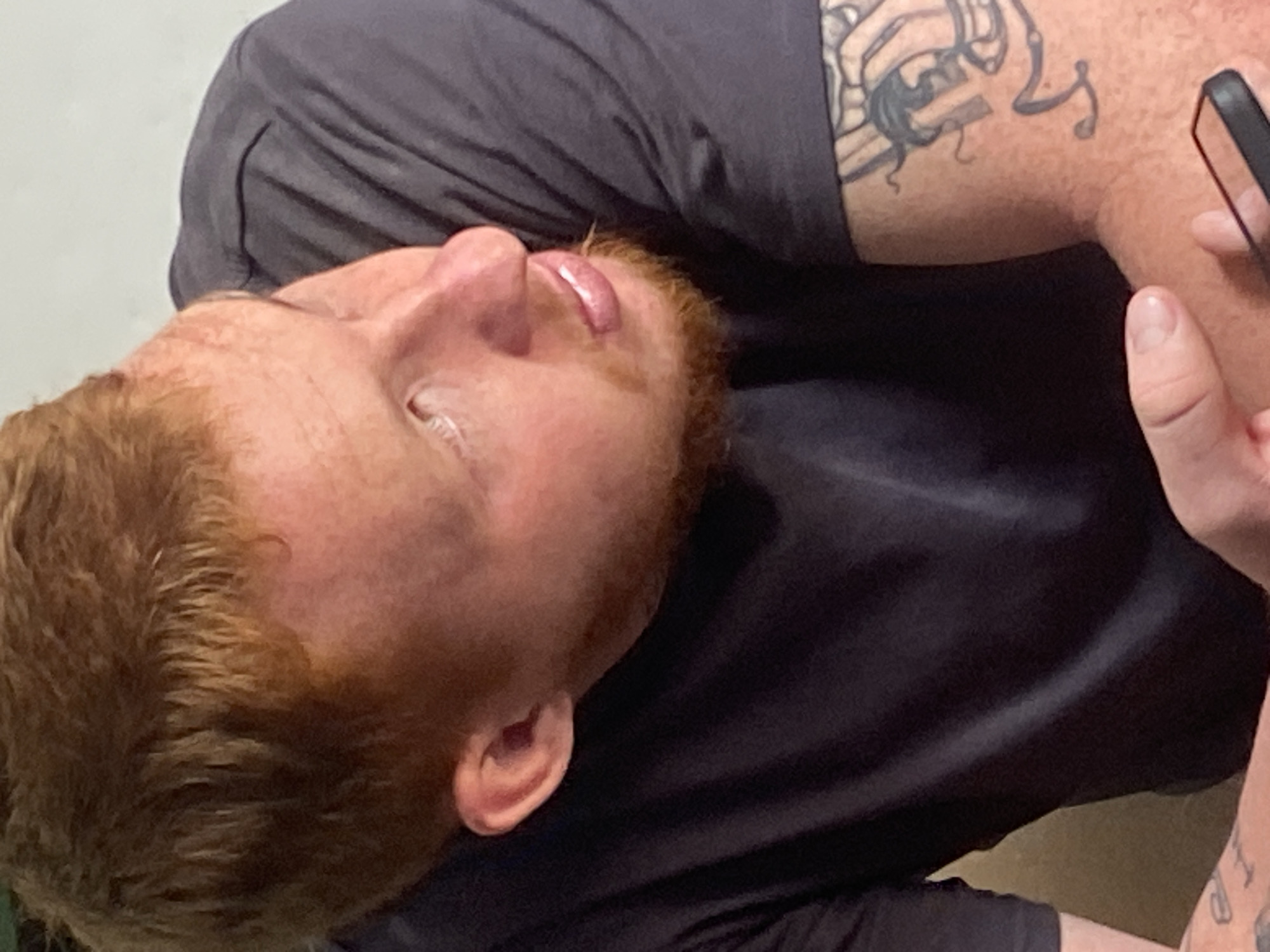 A man with red hair and a beard is shown from the chest up. He is wearing a dark grey t-shirt and has a tattoo on his left arm. His head is turned to the right, and he is looking upwards. His lips are slightly parted. He is holding a black object, possibly a phone, in his right hand. The background is a plain, light-colored wall.
