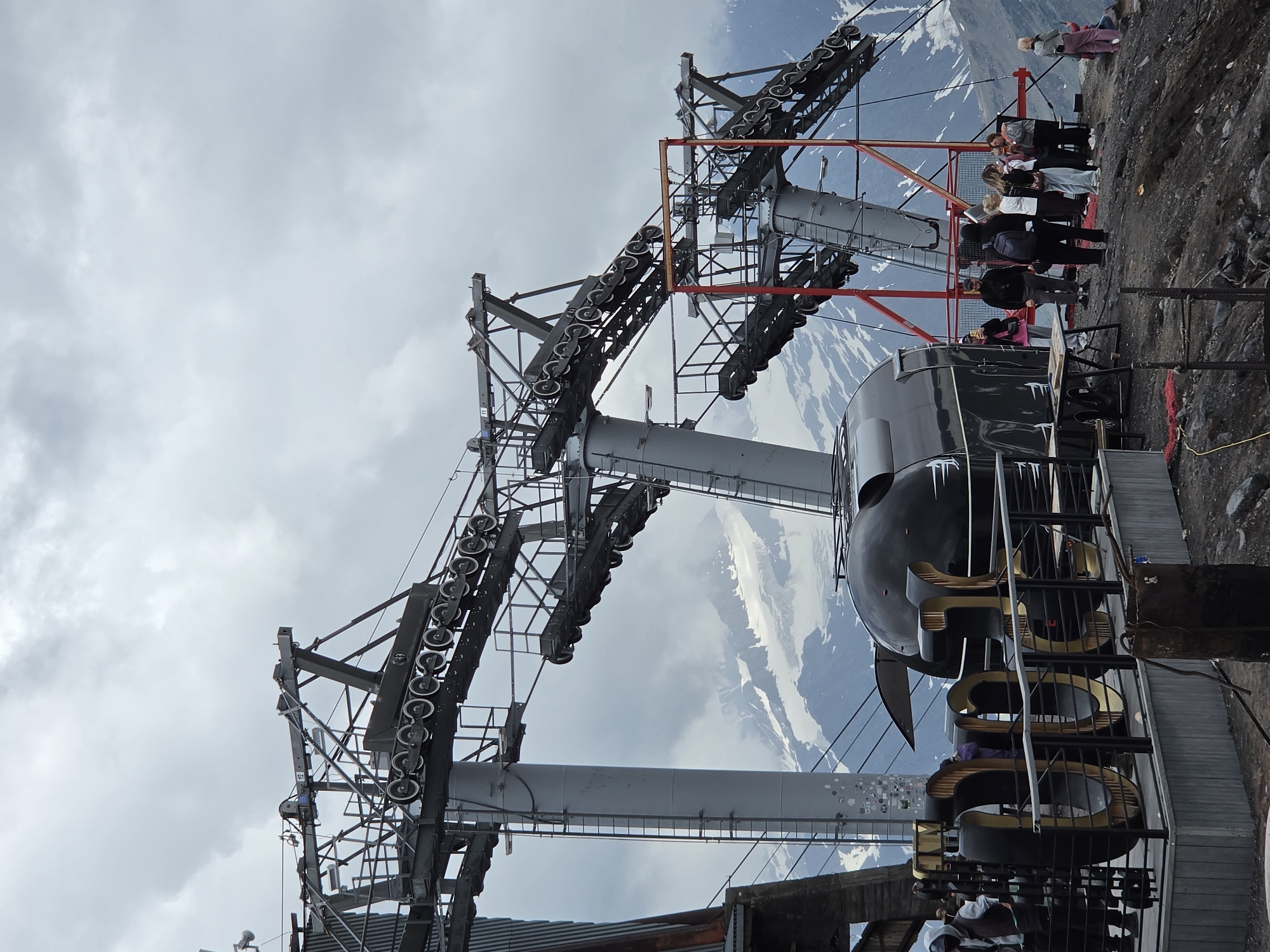 The image shows a ski lift station in Terskol, Russia, with snow-capped mountains in the background under a cloudy sky. The ski lift structure is prominent, featuring large metal supports and a series of pulleys for the cables. A large, black, oval-shaped cable car cabin is visible, along with large, golden, metallic letters spelling out "9000" in the foreground. Several people are gathered on a viewing platform, some taking pictures and others conversing. The ground is rocky and uneven, typical of a mountainous terrain.