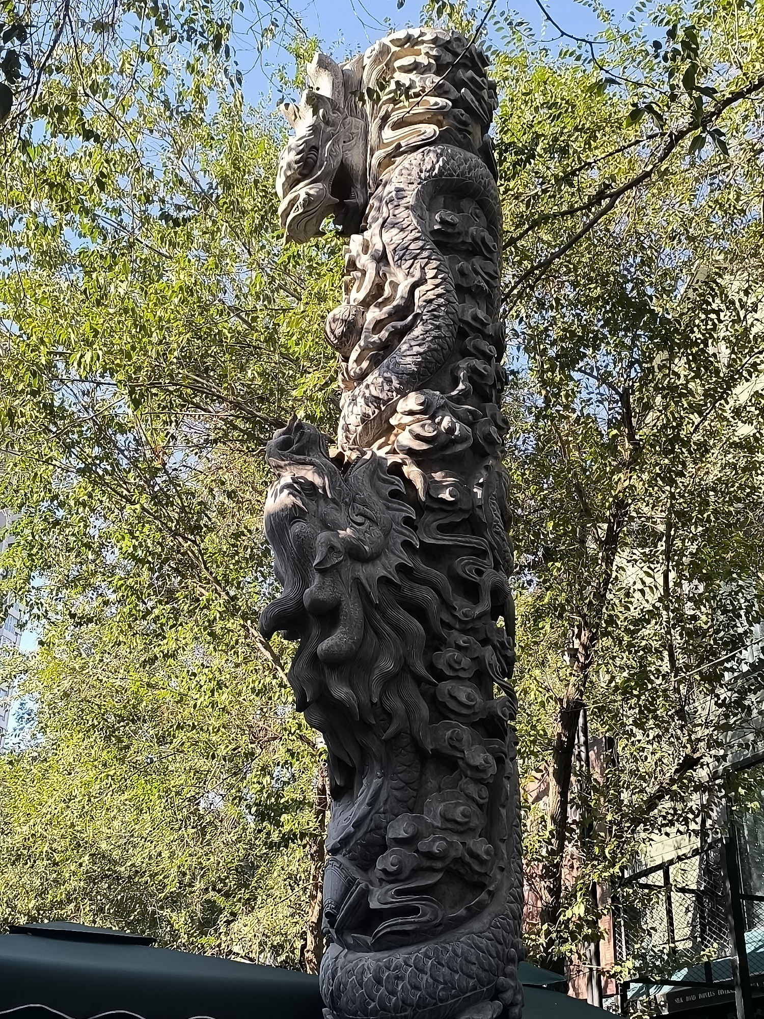 A stone column carved with intricate dragon motifs stands tall against a backdrop of lush green trees and a clear blue sky. The dragon's head is prominently featured at the top of the column, with its body coiling downwards, detailed with scales and flowing mane. The sculpture exhibits a weathered appearance, suggesting it is an outdoor installation that has been exposed to the elements. The surrounding trees are full of leaves, indicating a warm season. The bottom of the column is partially obscured by a dark green awning, and a metal fence is visible to the right, suggesting an urban setting. This statue is located in the city of Ürümqi, China.