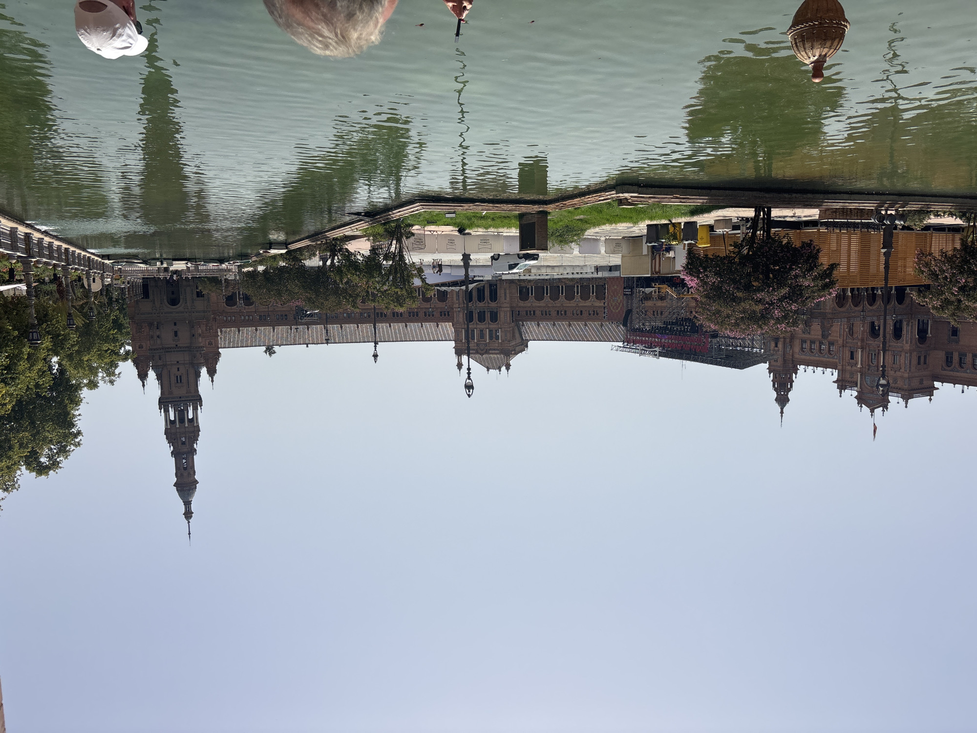 The image depicts a sunny day at Plaza de España in Seville, Spain, with a wide canal in the foreground reflecting the ornate architecture of the surrounding buildings. The central building is a large, reddish-brown structure with multiple arched windows and a tall, slender tower with a spire on the right side. Trees with pink blossoms are visible on the left, adjacent to temporary wooden structures and construction barriers. A white van and several people are present near the building. The canal has a serene surface with a few floating objects and a decorative element near the bottom left. In the foreground, two individuals are partially visible, one wearing a white baseball cap and looking towards the scene, the other holding a phone. The sky is clear and light blue, indicating fair weather.