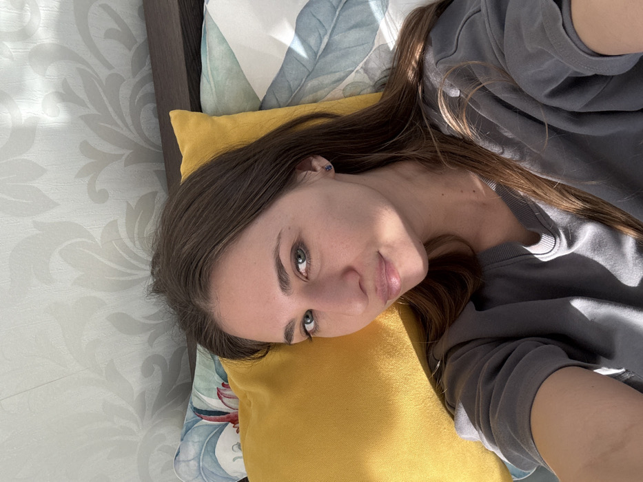 A close-up, eye-level indoor selfie depicts a female subject, appearing to be in her 20s to 30s, with long brown hair, light-colored eyes, and fair skin. She is positioned in a reclining posture, framed by a bright yellow pillow behind her head and a patterned pillow with a light blue and green botanical design to her right. The subject wears a plain dark grey long-sleeved v-neck top and a dark stud earring in her left ear. Her gaze is directed at the camera, and her lips are slightly parted in a faint smile. Direct sunlight from an unseen source casts distinct shadows across her face, right arm, and the pillows. The background reveals light-toned wallpaper with a subtle, repeating floral or damask pattern, and a dark wooden headboard partially visible behind the pillows. The specific location within the city of Saint Petersburg, Russia, cannot be determined from the visual evidence presented in the image.