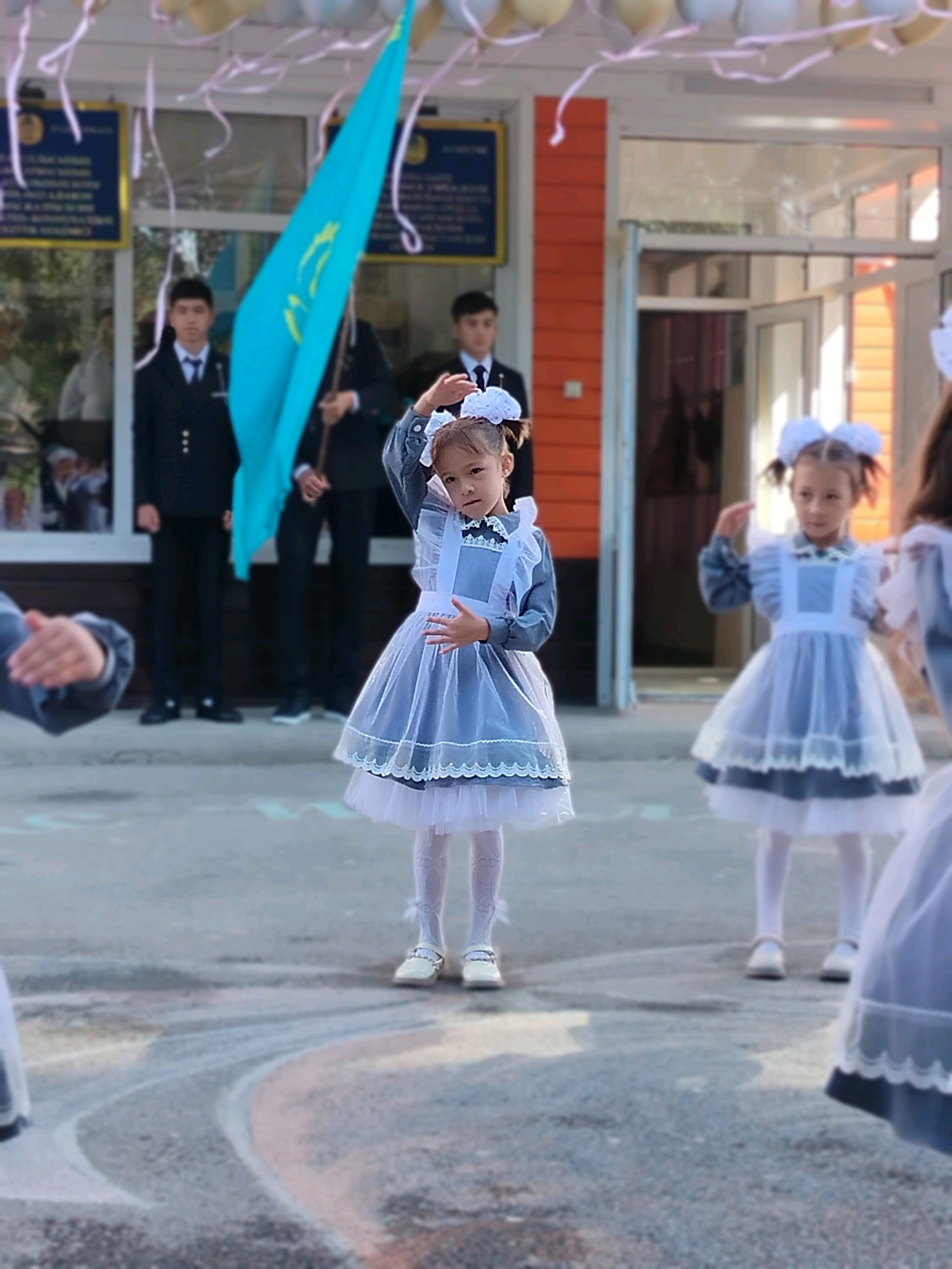 In the city of Kölkent, Kazakhstan, a group of schoolchildren are gathered outdoors, likely for an event or ceremony. In the foreground, a young girl in a traditional school uniform, complete with a white apron and large white hair bows, stands facing the camera with her right hand raised. She is wearing white tights and pale shoes. To her right, another girl in a similar uniform is also facing forward, with her left hand slightly raised. To the left, a blurry image of a child's outstretched hand is visible, also in a school uniform. In the background, several male students in dark uniforms and white shirts stand behind a large, bright turquoise flag featuring a golden sun and eagle. The backdrop features the orange-colored exterior of a building with large glass doors. Balloons and streamers are visible overhead, suggesting a festive occasion. The ground is paved and shows faint chalk markings.