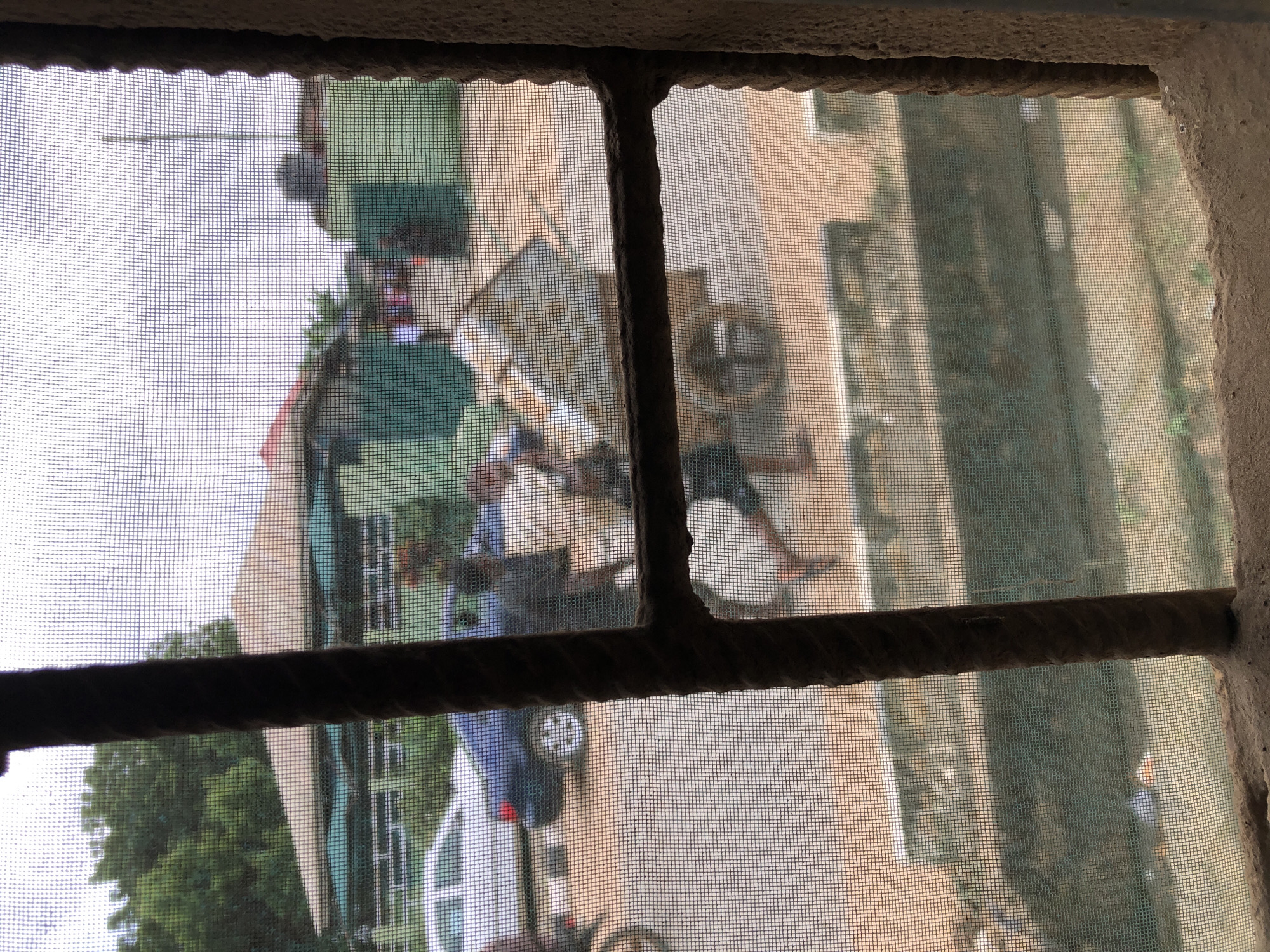 This image, taken through a window screen with a thick, rough frame, depicts a street scene in Lagos, Nigeria. The foreground is dominated by the window frame and screen, obscuring some details. In the mid-ground, a person is pushing a wheelbarrow across a dirt and paved road. Behind the wheelbarrow, a dark-colored car is parked. Further in the background, several buildings with distinctive green and brown facades are visible, along with trees. The sky is overcast. The scene suggests a typical day in a bustling urban environment, with people engaged in work and daily activities.