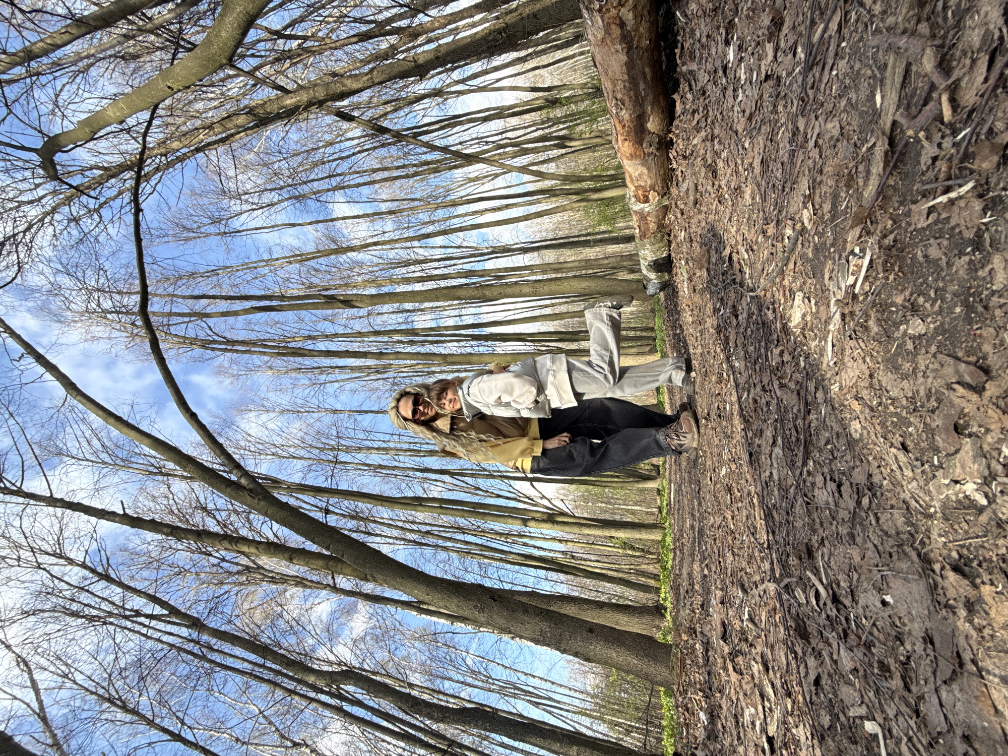 Two people are standing on a wooded path in the city of Rusanovo, Russia. The ground is covered with fallen leaves and branches, and tall, leafless trees line the path on either side. A bright blue sky is visible through the trees. One person is wearing a mustard-colored jacket, the other a light gray jacket. They are embracing and looking at the camera.