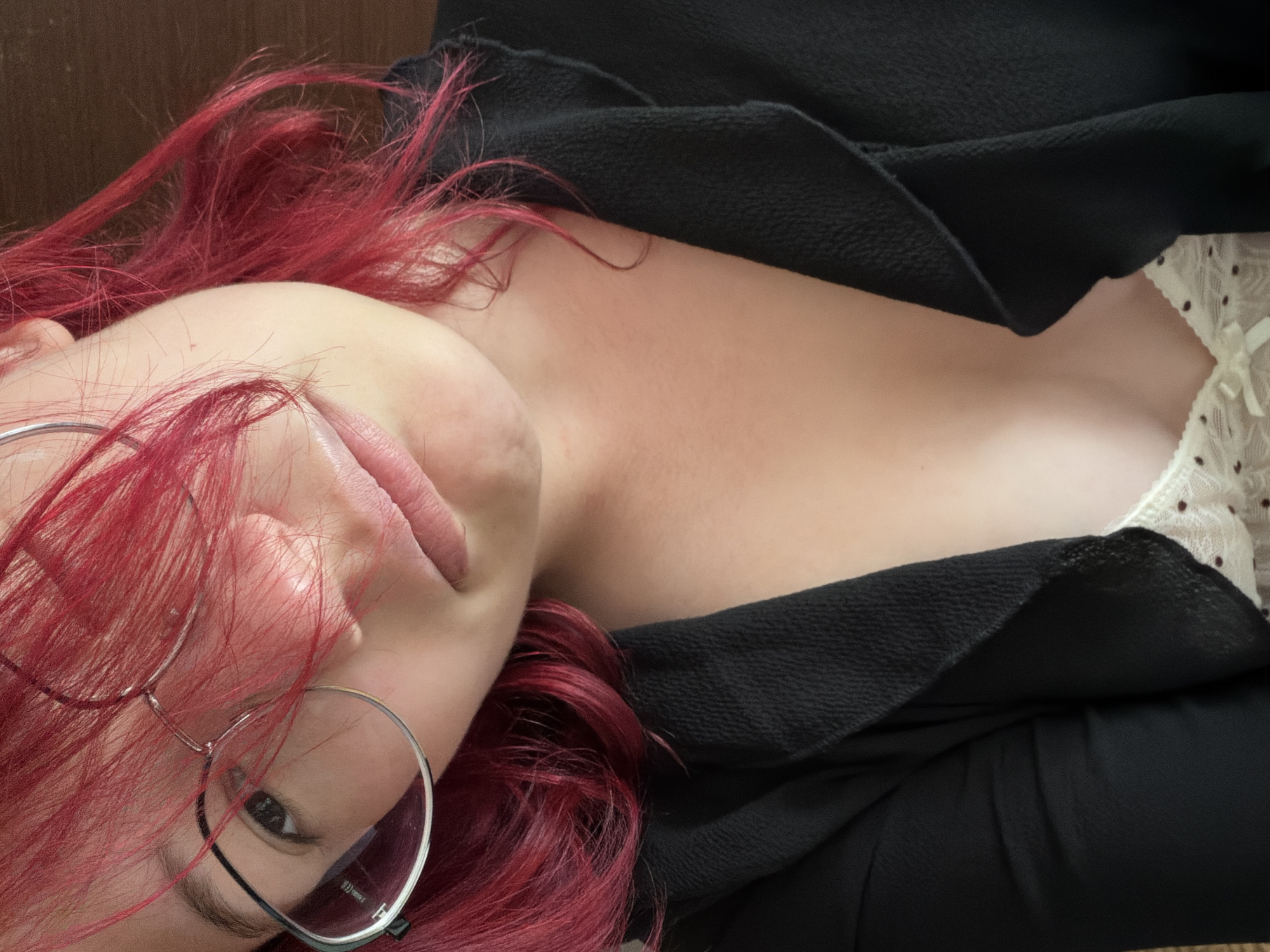 The image is a close-up selfie of a young person with bright red hair, wearing glasses and a black jacket over a white top with a polka-dot pattern. The person is looking towards the camera with a slight smile. The background is a plain wooden surface. There is no indication of the location being in Chelyabinsk, Russia.