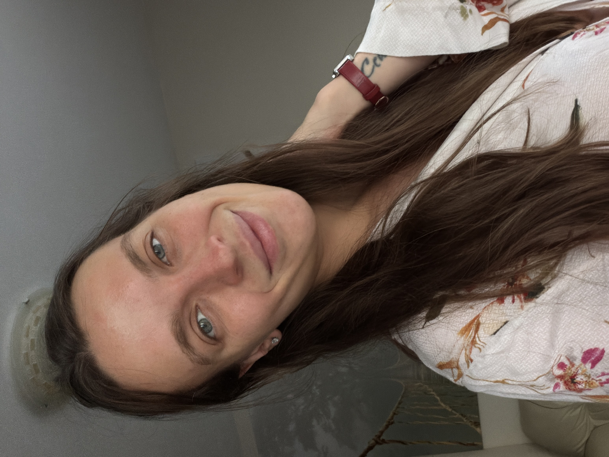 A woman with long brown hair and blue eyes is facing the camera. She is wearing a white floral shirt and a red watch on her left wrist. A tattoo is visible on her left arm. The background consists of a gray wall and what appears to be a blurred, light-colored object or furniture. The scene lacks enough information to determine the specific location in Budva, Montenegro.