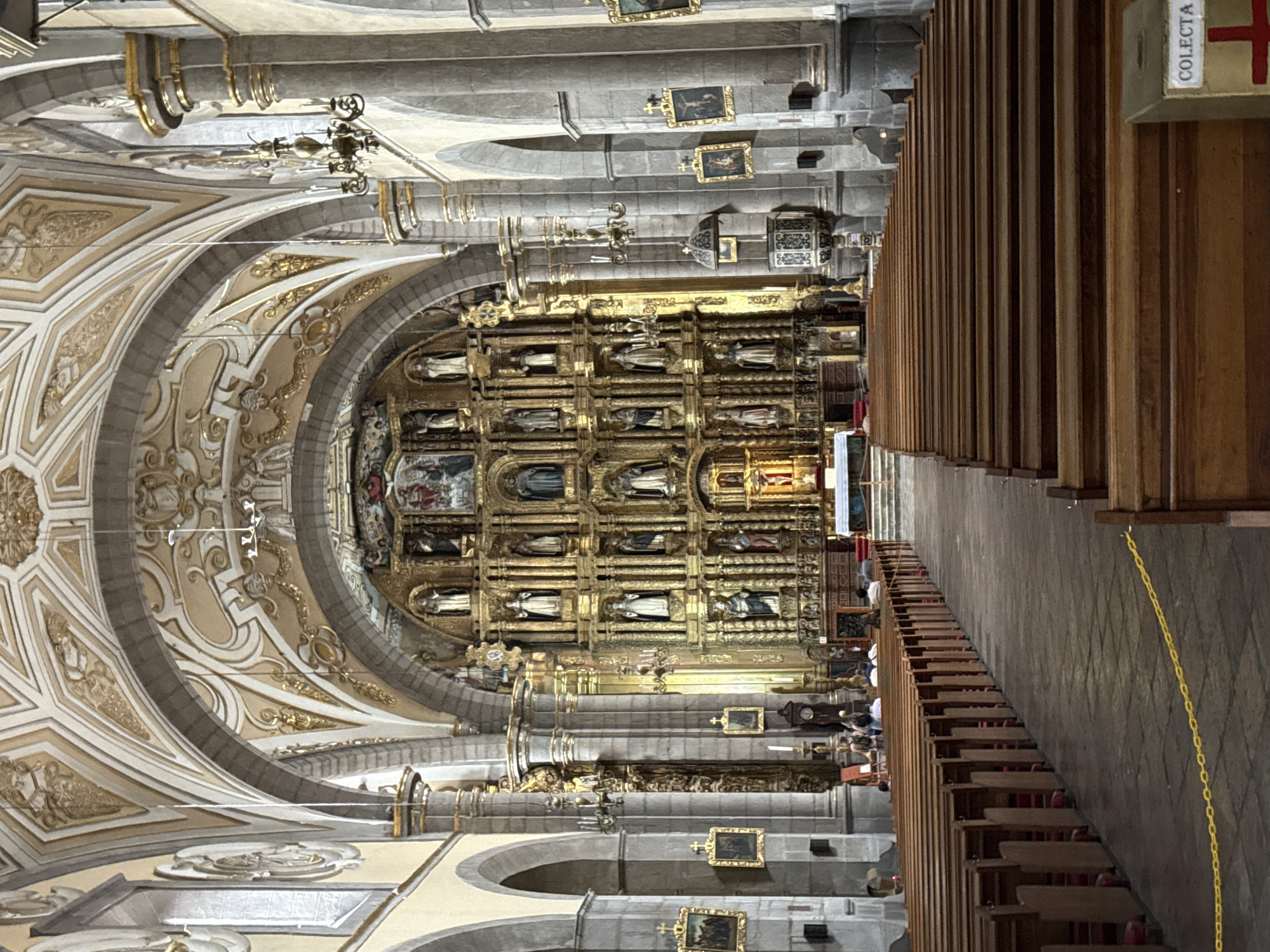 The image displays the ornate interior of a grand church or cathedral, viewed from a side aisle looking towards the main altar. The photograph is oriented vertically, rotated 90 degrees clockwise.
The scene is dominated by elaborate Baroque or Churrigueresque architecture. High, vaulted ceilings feature intricate stucco work in white and gold, forming decorative arches and patterns. Supporting these are robust stone columns and arches.
At the far end, a towering, multi-tiered gilded altarpiece (retablo) serves as the focal point. This golden structure is richly carved and houses numerous statues of saints and religious figures within individual niches, arranged in multiple horizontal and vertical sections. Some small paintings are also integrated into the altarpiece and adorn the stone walls between the architectural bays.
In the foreground and midground, rows of dark wooden pews line the side aisle. Hanging from the ceiling are ornate chandeliers or lighting fixtures. Attached to the end of a pew in the upper right corner is a wooden collection box, labeled "COLECTA" (Collection/Offering) next to a red cross. A yellow chain runs along the edge of the pews.
Several people are visible in the distance near the altar and seated in the pews, though their faces are indistinct. The activity appears to be quiet contemplation or visitation. The church's opulent style and historical grandeur are consistent with the rich Spanish colonial architecture found in Puebla, Mexico. The lighting suggests daytime.