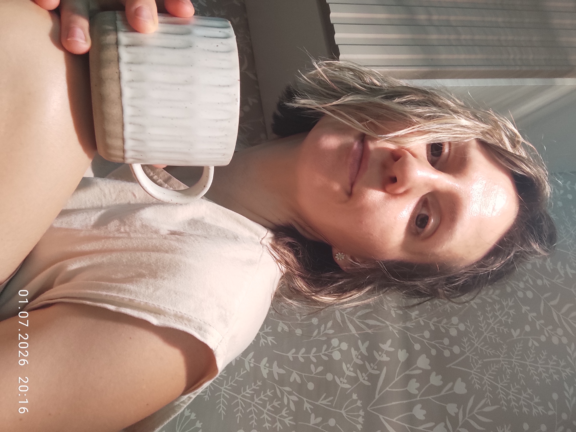 The image shows a person in a close-up, angled shot, holding a mug. The subject is lying down, with their head tilted back and their face partially obscured by their hand holding the mug. Sunlight is streaming in from the right, creating highlights on their skin and hair, and casting shadows across the scene. The background appears to be a bed with a patterned duvet cover. The person is wearing a light-colored t-shirt. Their expression is relaxed, with their eyes open and looking towards the camera.
Visible text on the image reads: "01.07.2026 20:16". This indicates the date and time the photo was taken. The lighting suggests it is late afternoon or early evening, consistent with the time stamp. There are no other discernible location cues in the image itself, but the provided context places the location as Malyi Bukor, Russia.