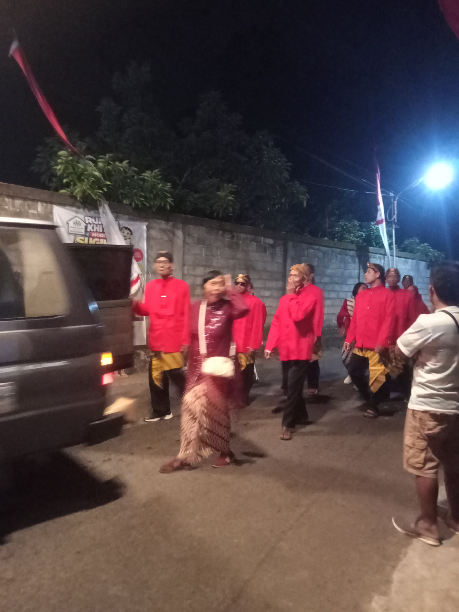 In Urutsewu, Indonesia, at night, a group of people in traditional attire walk down a street lined with a brick wall and trees. The individuals are wearing red tops and patterned lower garments, suggesting a ceremonial or cultural event. A dark-colored van is partially visible on the left side of the image, with its taillight illuminated. A street lamp is visible on the right, casting light on the scene. A flagpole with a red and white flag, likely the Indonesian flag, is also present. The ground is paved and appears wet, indicating recent rain. The overall atmosphere is dark, illuminated by artificial lights and a bright street lamp, creating a somewhat hazy effect.