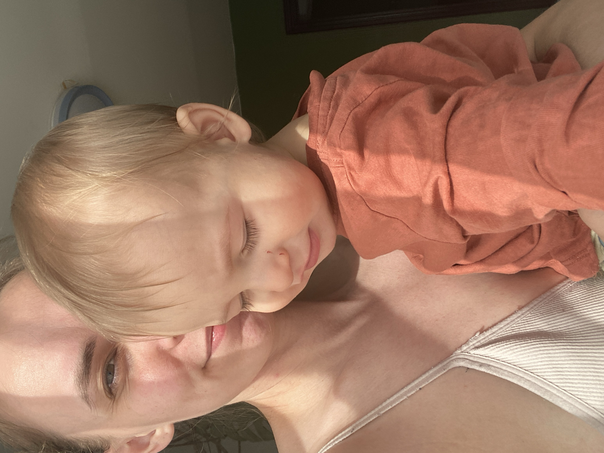 A close-up, slightly rotated, indoor shot depicts a woman with light skin and a child, likely a toddler, lying on her chest. The child, dressed in a rust-colored shirt, has their eyes closed and is asleep or resting peacefully. The woman's face is partially visible, with one eye open. The lighting is uneven, casting shadows across their faces. A white tank top is visible on the woman. In the background, out of focus, are hints of a wall, possibly green in color, and a circular object on the ceiling. The location, Chaikovsky, Russia, is not discernible from the image.