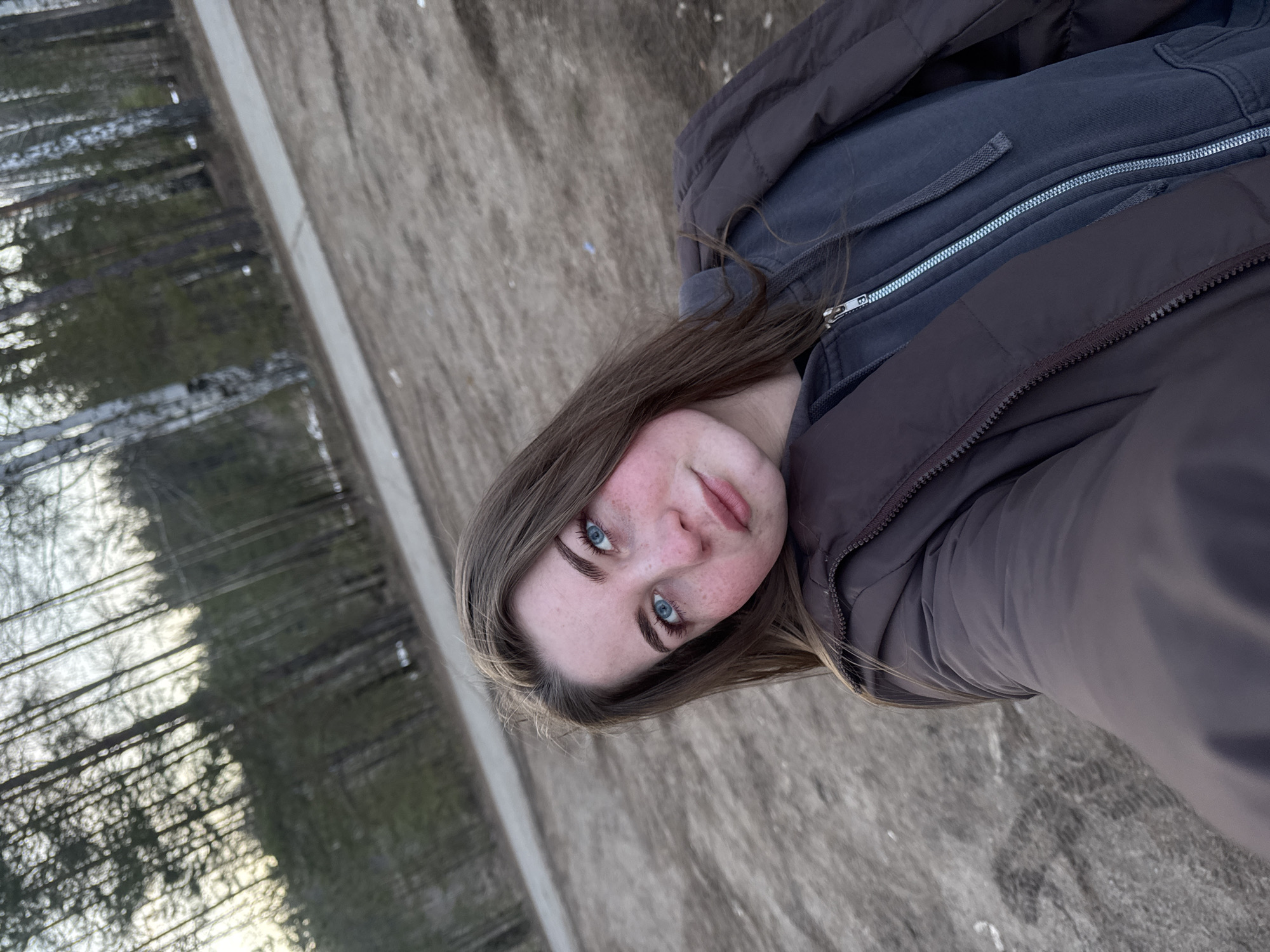 A portrait of a young woman with light brown hair and blue eyes, taken outdoors. She is wearing a brown jacket over a gray zippered hoodie. The background consists of a dirt path with a line of trees visible on the left side of the image. The photo may have been taken in a park or natural area, possibly in Noviy, Russia. The image is rotated clockwise.