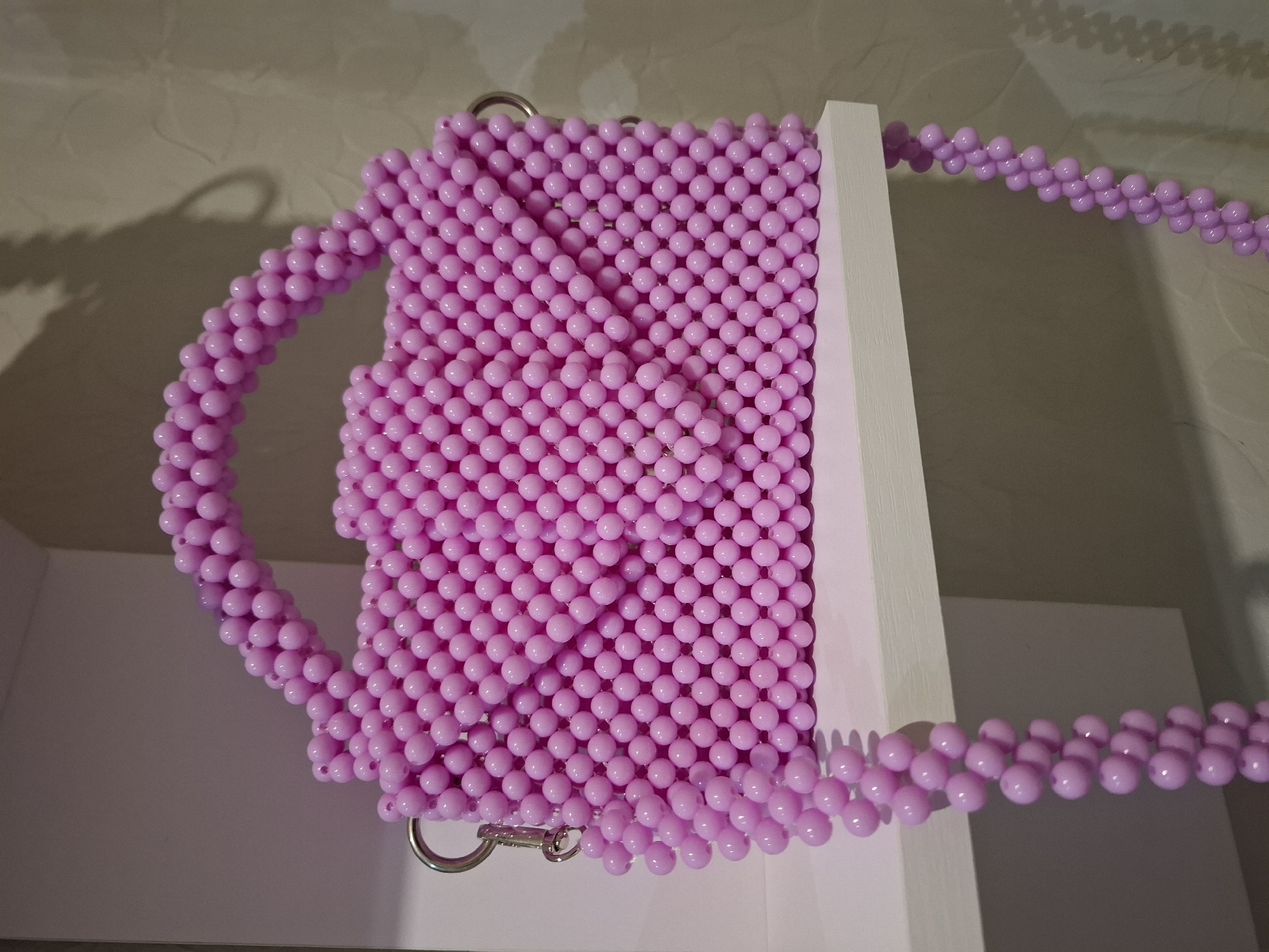 A close-up, eye-level shot shows a pink beaded handbag resting on a white shelf. The handbag has a structured shape with a flap closure and a top handle, also made of beads. A beaded strap hangs down the side of the bag, extending off the shelf. The background consists of a textured wall, suggesting an indoor setting. The image does not contain any elements that would allow for a determination of its location in Korenovsk, Russia.