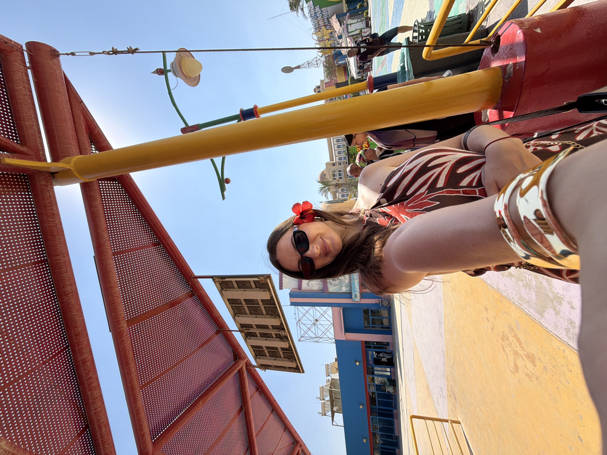 In Ash Sheikh Zayed, Egypt, a woman with dark hair and sunglasses takes a selfie outdoors. She is wearing a brown and white patterned dress and a red flower in her hair. She is also wearing a gold bracelet. The background shows a colorful outdoor space with a red canopy, yellow poles, and buildings in the distance. There are other people in the background, and some solar water heaters are visible on one of the buildings. The ground is paved with yellow and pink concrete.