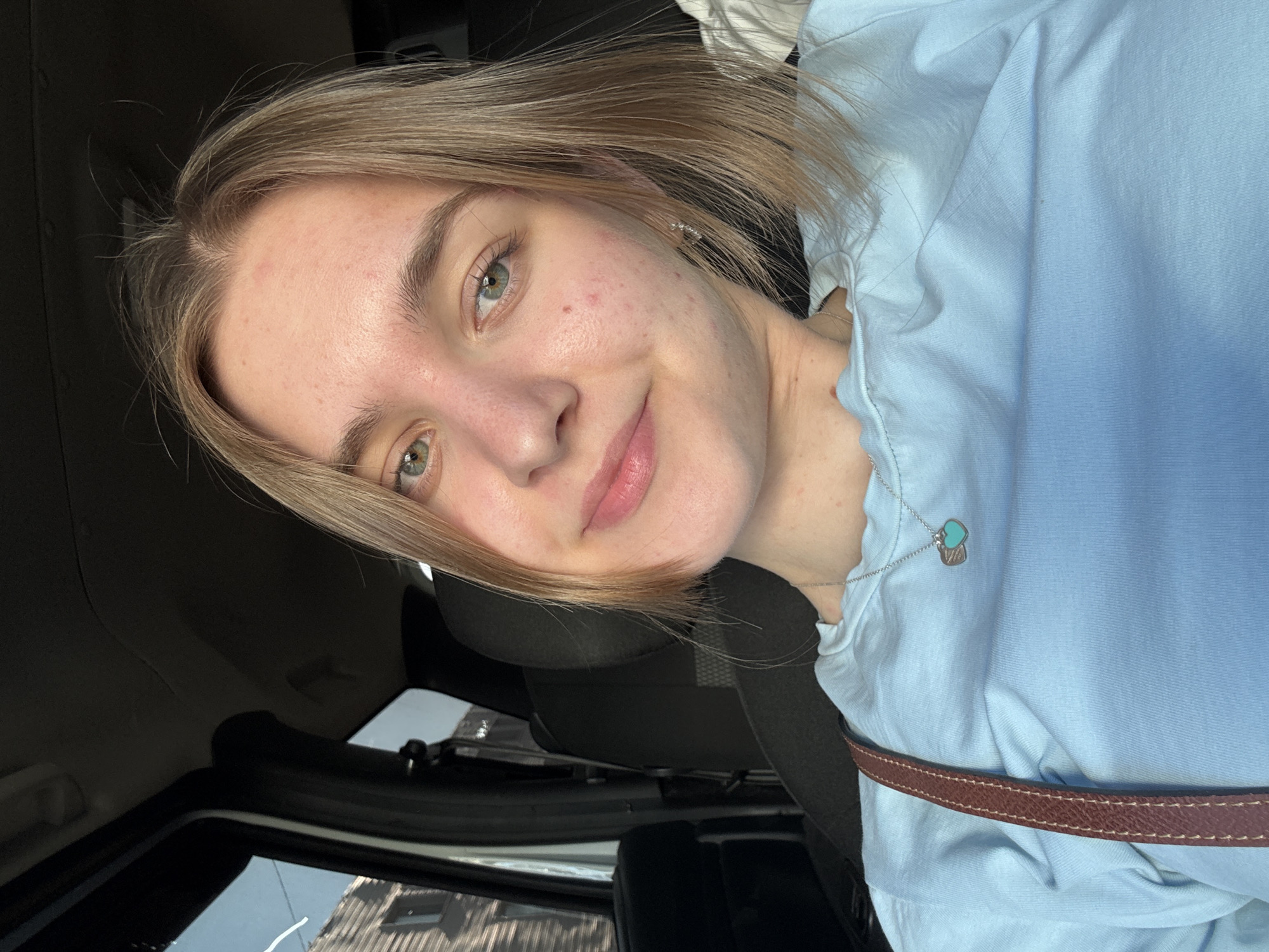 A selfie of a young woman with short, blonde hair and light skin. She is wearing a light blue shirt and a necklace with a turquoise pendant. She is inside a car with dark upholstery, possibly in Perm, Russia, based on visual analysis of exterior elements and the camera's location settings. A brown strap, possibly a seatbelt, is visible across her shirt. The woman is smiling slightly at the camera. The view through the car window shows a building with multiple windows.