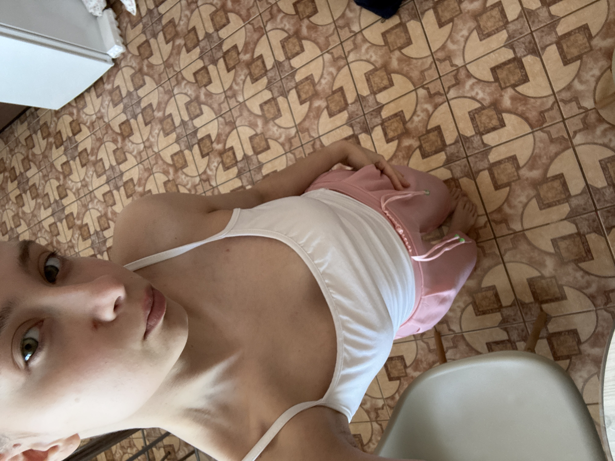 A young individual with fair skin and green eyes is shown in a close-up, angled shot. They are wearing a white tank top and pink bottoms. The location appears to be indoors, as suggested by the tiled floor with a geometric pattern. A white cabinet is visible in the top left of the frame, and a pale gray chair is in the bottom right. The individual's expression is neutral as they look directly at the camera. The image was taken in Прикамский, Russia.