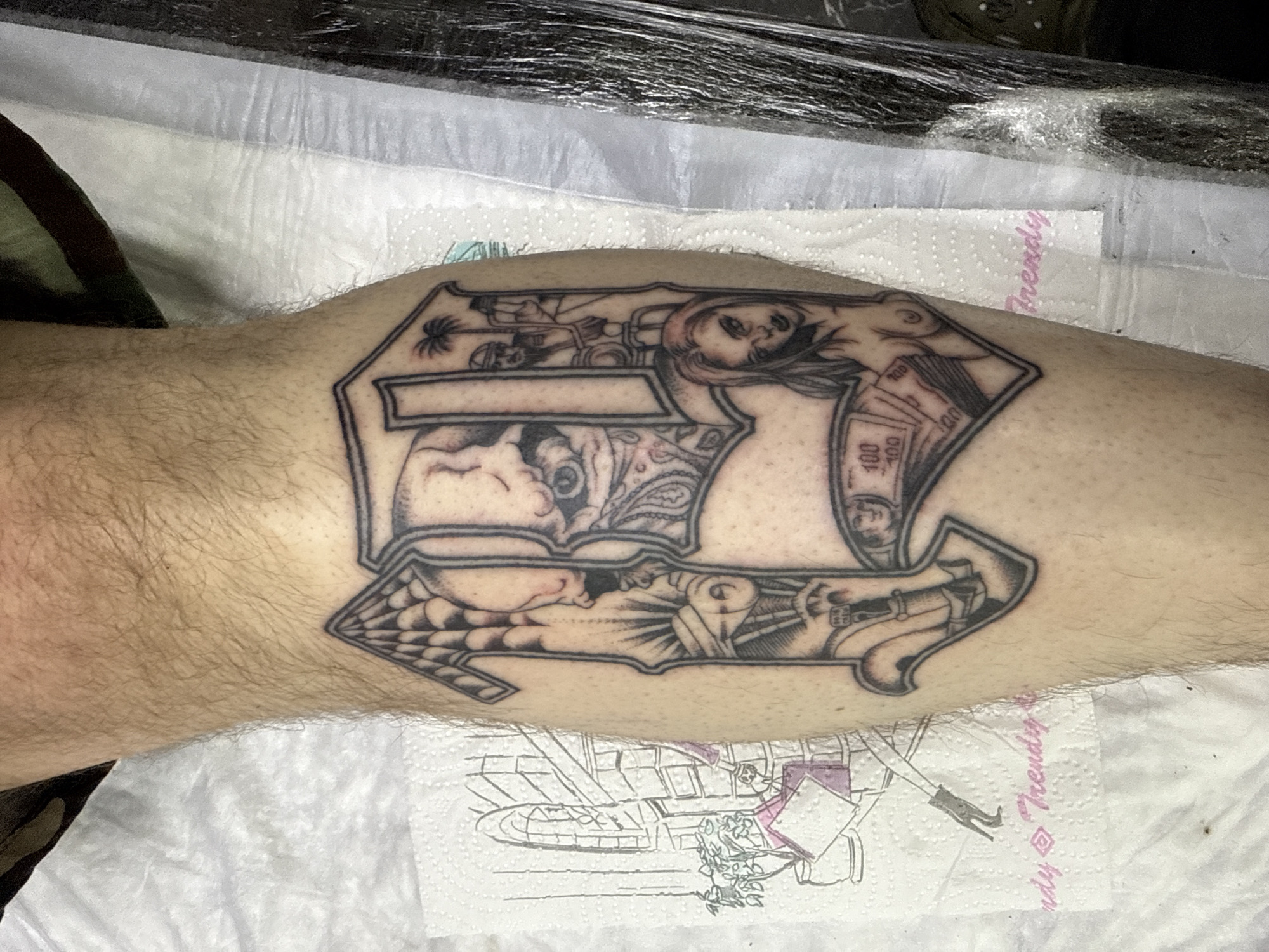 A close-up, high-angle shot shows a tattoo on a person's lower leg. The tattoo is primarily in black ink with a grayscale appearance. The tattoo's design includes multiple scenes or images:
- At the top, there's a woman's face, and what appears to be a stack of money. There is a palm tree, what looks like a soldier, and a blank box.
- At the bottom, the tattoo design includes a sleeping figure and a pistol. The tattoo has a cobweb.
The leg has fine hair. The leg rests on a white surface, possibly paper towels. One paper towel has writing on it.
The picture does not contain the city of Moscow.