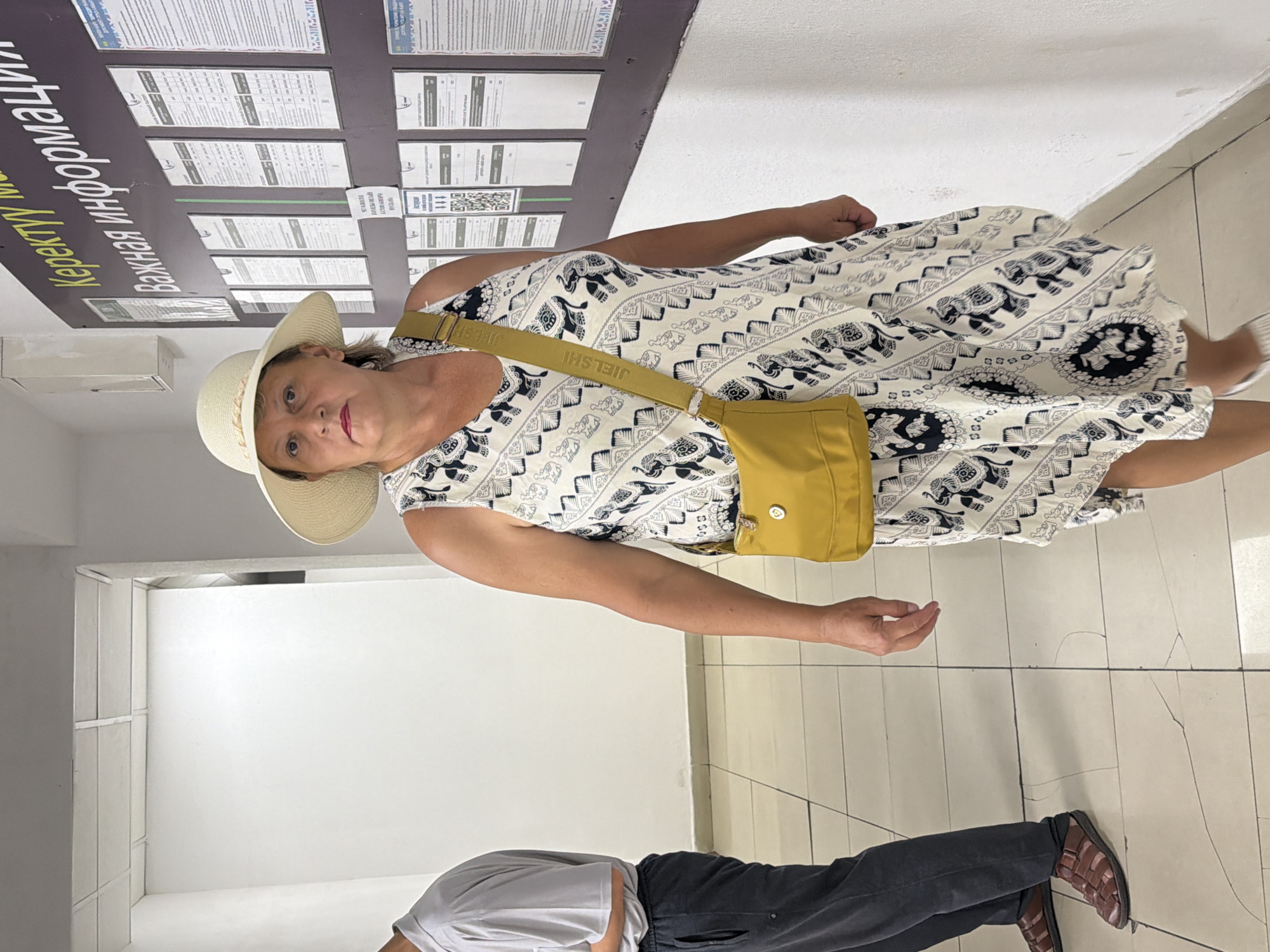 A woman wearing a white wide-brimmed hat and a sleeveless white dress with a black elephant pattern stands in an indoor hallway. The dress has a fitted bodice and flares out at the hem. A mustard-yellow crossbody bag with a black strap is slung over her right shoulder. The strap has black lettering. She looks directly at the camera with a neutral expression. To her left, a person wearing a gray polo shirt and dark pants stands with their back mostly to the camera, facing a white wall. They are wearing brown sandals. The hallway has a tiled floor with visible cracks and a white wall on the left. A dark bulletin board with multiple documents and notices is mounted on the wall to the right of the woman. The bulletin board has text in a foreign language, some of which is in yellow and white font. The ceiling is white with visible panels.