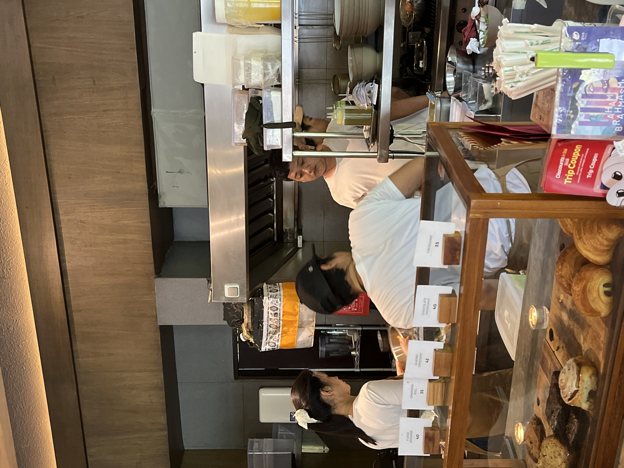 This image captures an interior scene, likely a cafe or bakery kitchen and serving area, located in Kuta Selatan, Indonesia. Three individuals, presumably staff, are visible working behind a counter.
In the foreground, a woman with dark hair tied back and a white bow, wearing a white t-shirt, stands facing right, looking towards a glass display case filled with pastries. The display case holds various baked goods, including croissants, brownies, and cinnamon rolls. Labels with prices are visible for these items.
Behind the counter, a man in a white t-shirt and dark hair is seen focused on an activity in the kitchen area, which features stainless steel shelving, various containers, and cooking equipment. Another person, largely obscured and wearing a dark cap and white t-shirt, is also working in the kitchen space, with their back partially to the viewer.
The setting suggests a bustling environment, with stacked items and food preparation equipment visible. On the customer-facing side of the counter, brochures and signs are displayed, some featuring text such as "Discounts in Bali," "Trip Coupon" (repeated), and "BRAHMASM." A bundle of straws, possibly reusable, is also present.
Visible text on the food labels includes:
* FUDGE BROWNIE 40
* CINNAMON ROLL 35
* ALMOND CROISSANT 42
* CHOCOLATE CROISSANT 40
* CROISSANT 35
The lighting appears bright and natural, indicating it is likely daytime. The overall atmosphere is one of focused work in a commercial food establishment.