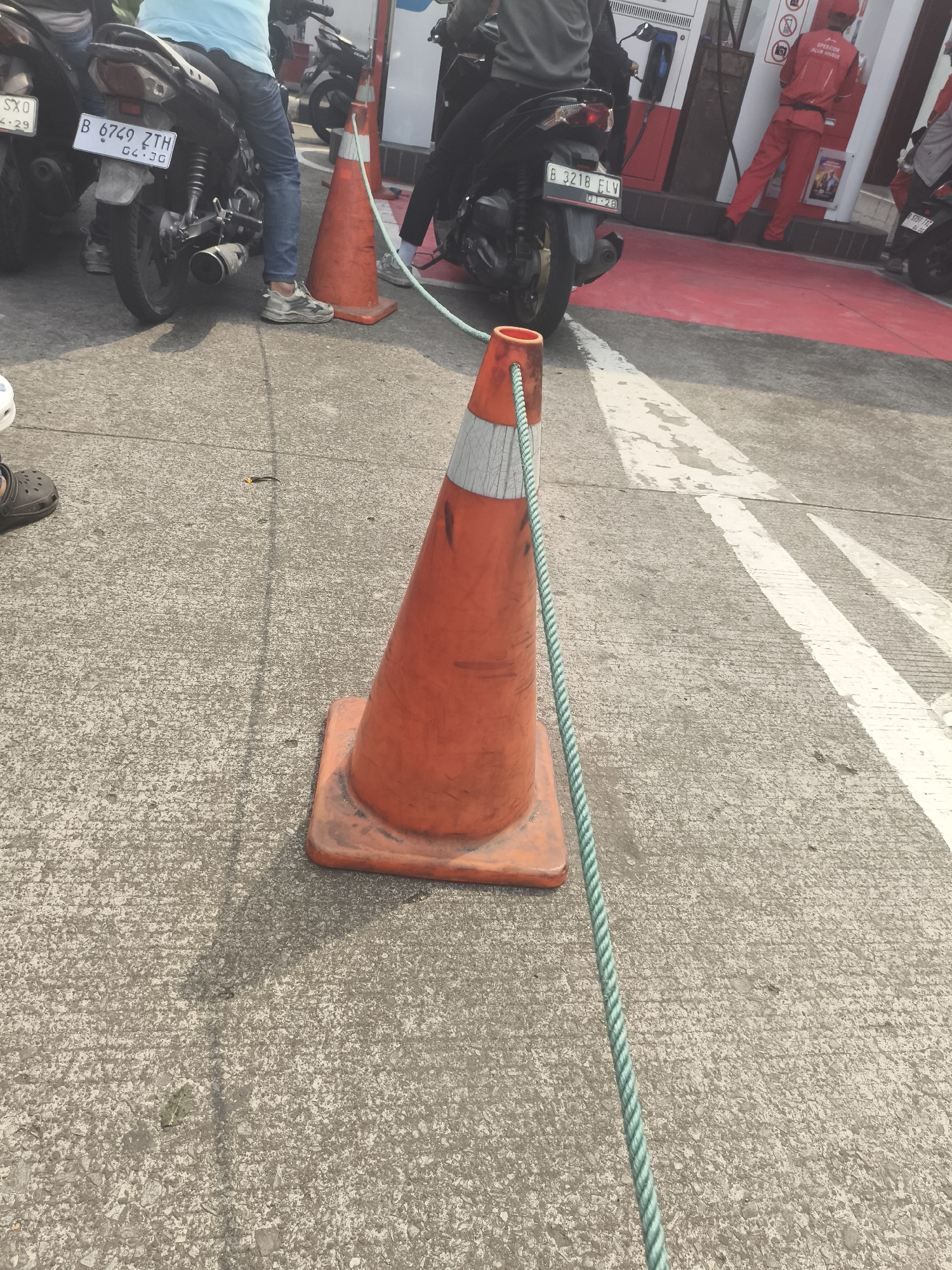 This image depicts a scene at a Pertamina gas station in Depok, Indonesia. In the foreground, an orange traffic cone with a green rope attached is prominently displayed. The cone appears weathered and has reflective tape. The ground is paved concrete with white painted lines. In the background, several motorcycles are parked, and people are visible. One person is wearing a light blue shirt and jeans, sitting on a motorcycle. Another person in the background is wearing a red uniform with "OPERATOR" visible on it, suggesting they are an attendant. The presence of motorcycles and a gas station strongly indicates this is a public space, likely a busy area given the context of a gas station.