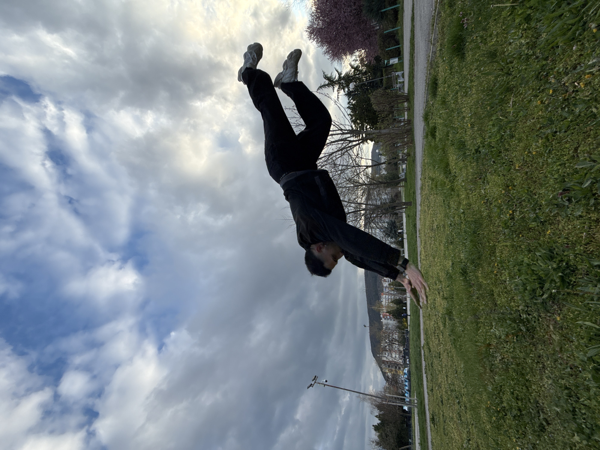 A person is performing a backflip on a grassy field in Shumen, Bulgaria. The person is wearing black pants, grey shoes, and a black jacket. The sky is cloudy. In the background, there is a city with buildings and trees. There is a grassy field and trees on the right side of the image. There is a road and street lights visible.