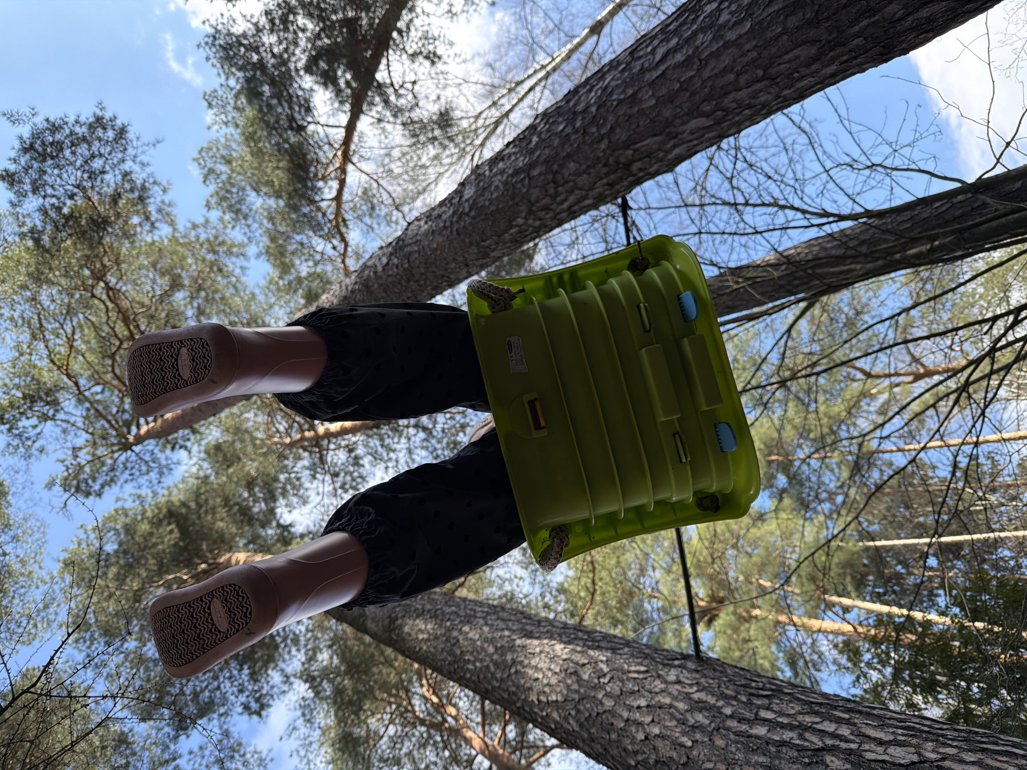 The image, taken from a low-angle perspective in an outdoor setting, possibly Udelnaya, Russia, depicts a green child's swing suspended between tree trunks. A pair of legs in dark, patterned pants and brown, textured-sole shoes are visible on the swing, appearing to be a doll or mannequin. The view extends upwards to include tree branches and a patch of blue sky.