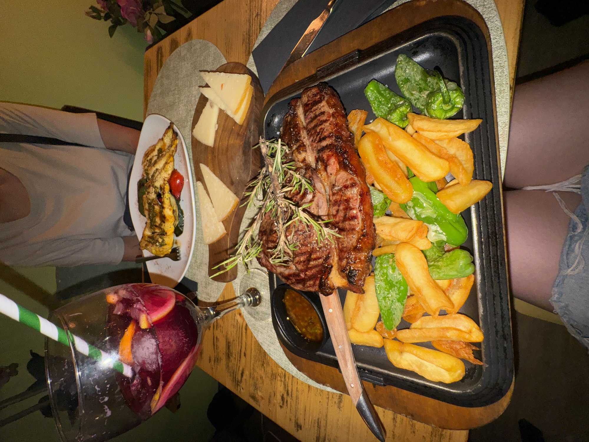 A tabletop scene features a meal with several components. A grilled steak adorned with rosemary is centrally placed on a black tray alongside thick-cut fries and blistered padron peppers. Next to the tray is a wooden board with slices of cheese. A white plate contains grilled vegetables, including zucchini, and a red tomato. A large glass of sangria with a green and white striped straw is in the foreground. Two individuals are partially visible; one on the left wearing a beige shirt, and another on the right in denim shorts. The table surface has a light brown, possibly wooden, finish. The setting is dimly lit. The location is inferred to be Barcelona, Spain.