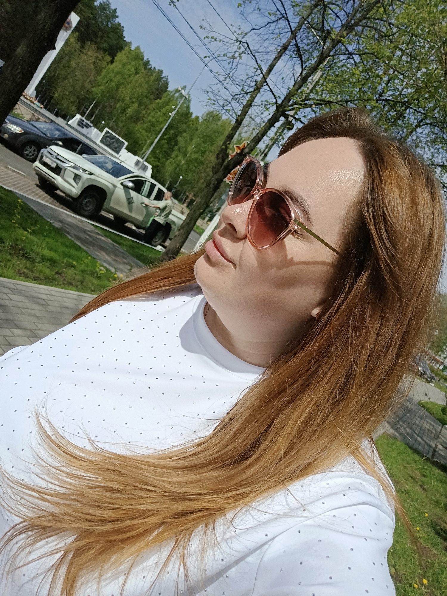 A woman with long, light brown hair is wearing pink sunglasses and a white, dotted shirt outdoors. A black sedan and a white pickup truck are visible in the background, parked on a road next to a green grassy area and trees. The woman is in Chaikovsky, Russia.